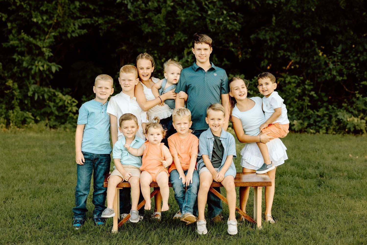 Group of eleven children posing together on grass for a family portrait in a lush green outdoor setting.