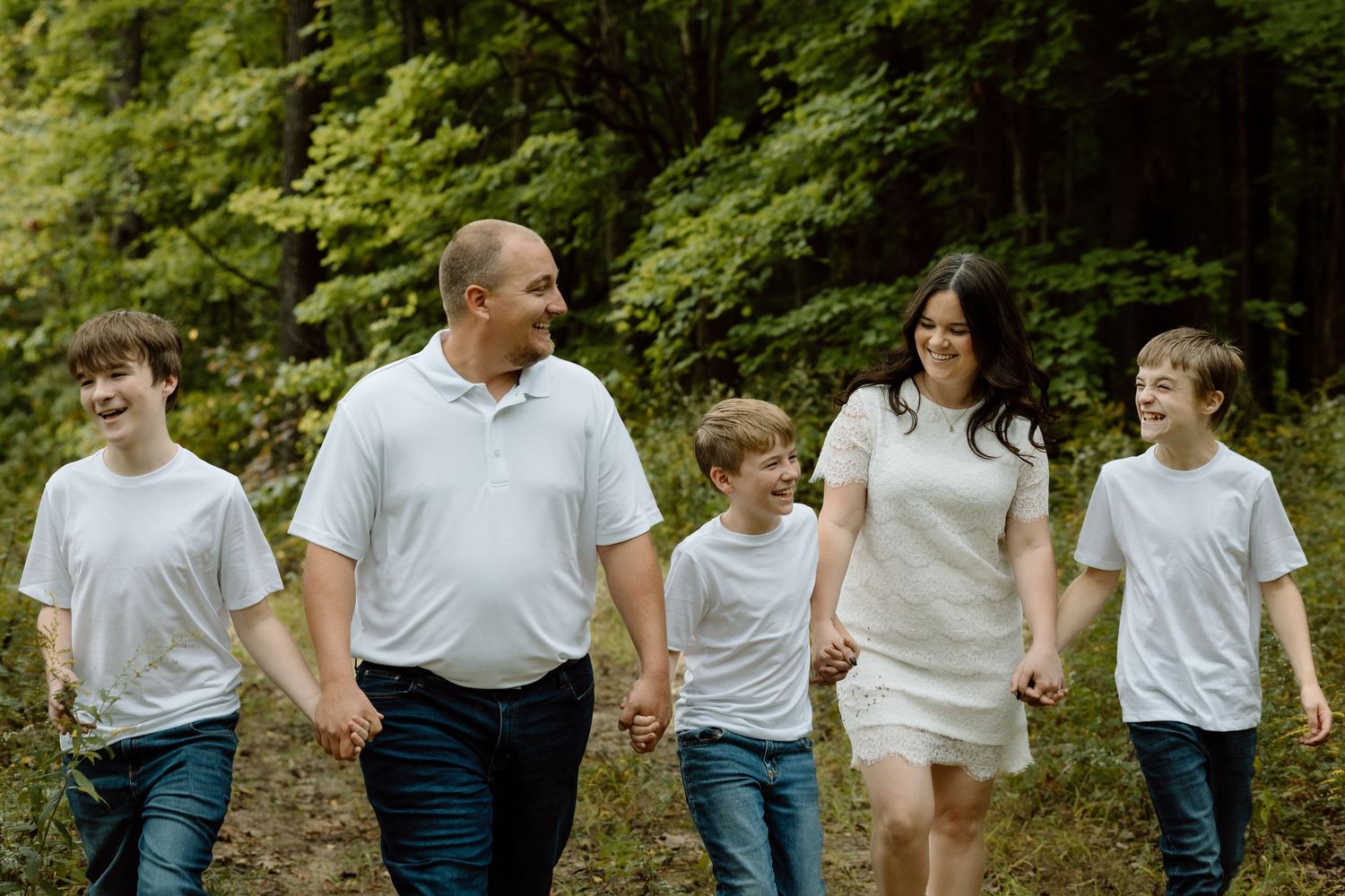 Happy family of five walking hand-in-hand along a wooded path in matching white outfits.