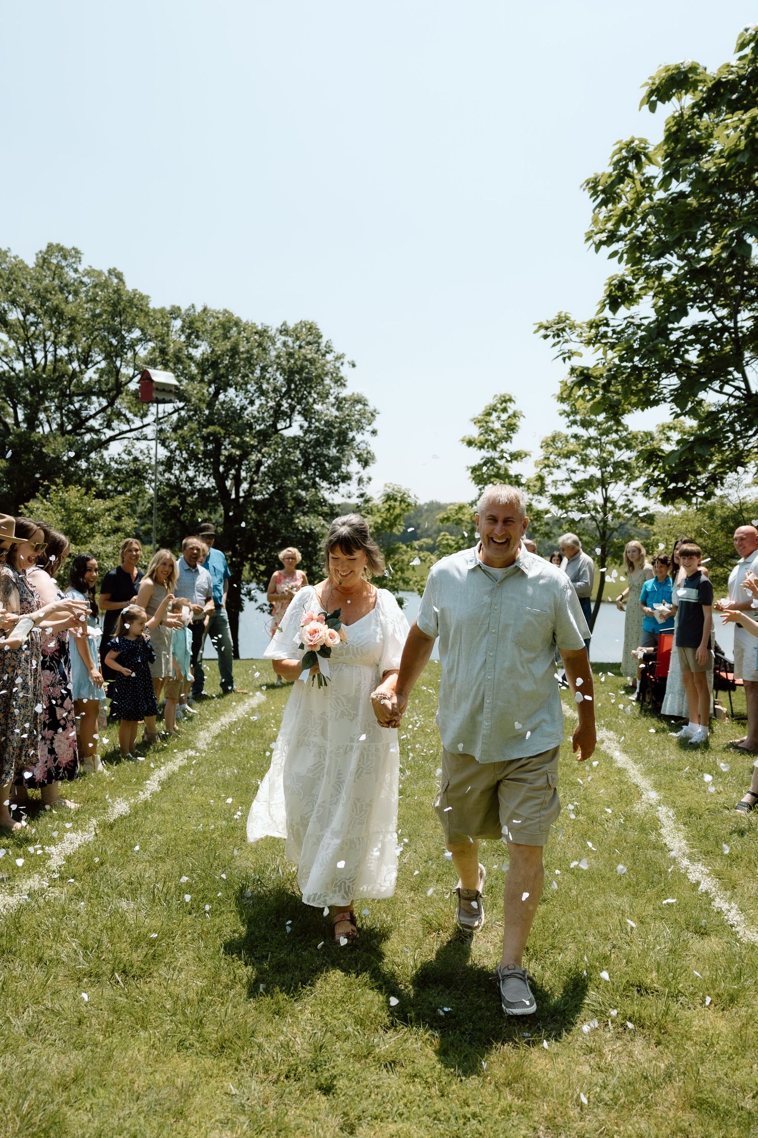 Newlywed couple walks through confetti-throwing guests after outdoor lakeside wedding ceremony.