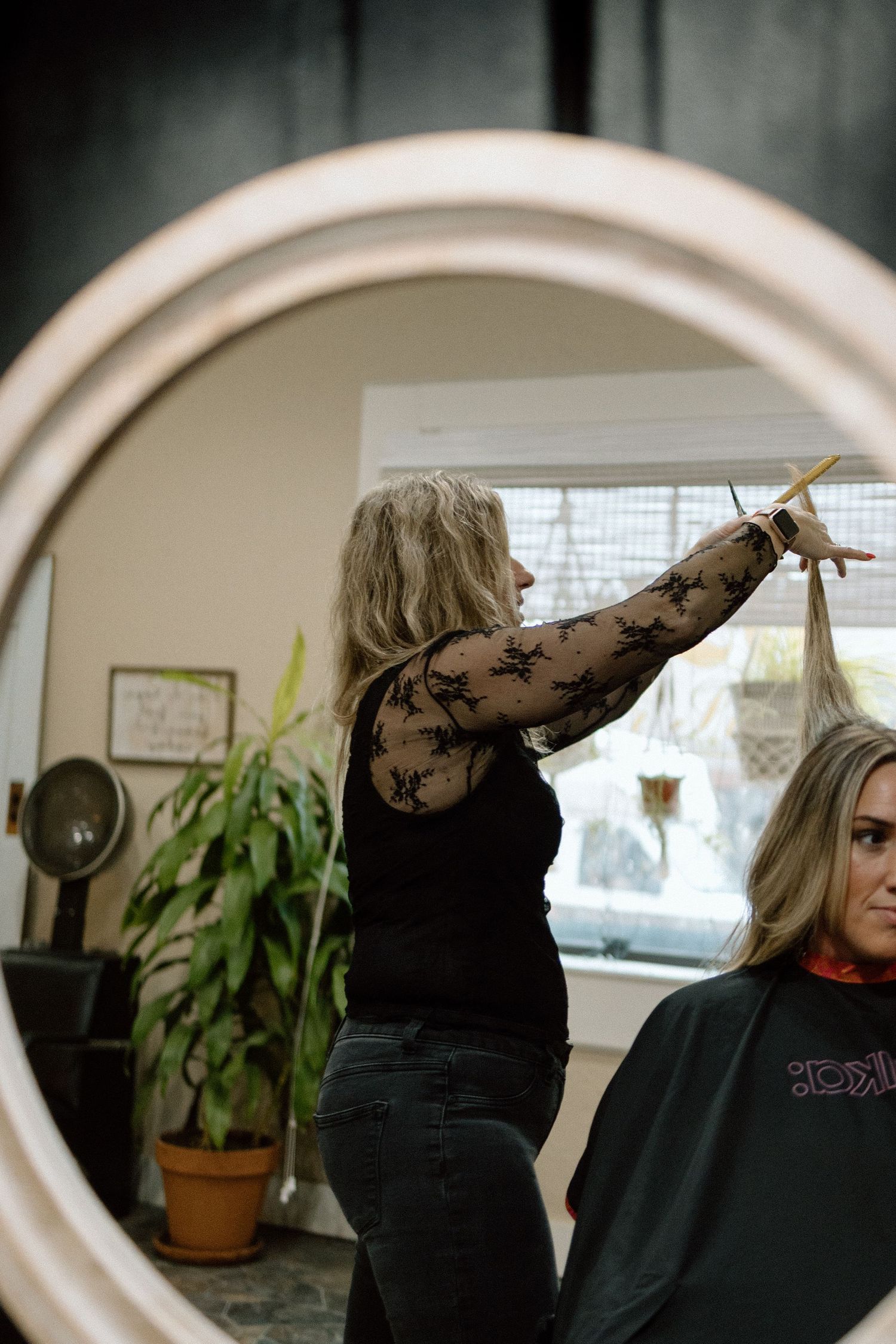 A hairstylist cutting a client's hair, reflected in a round mirror in a salon setting.