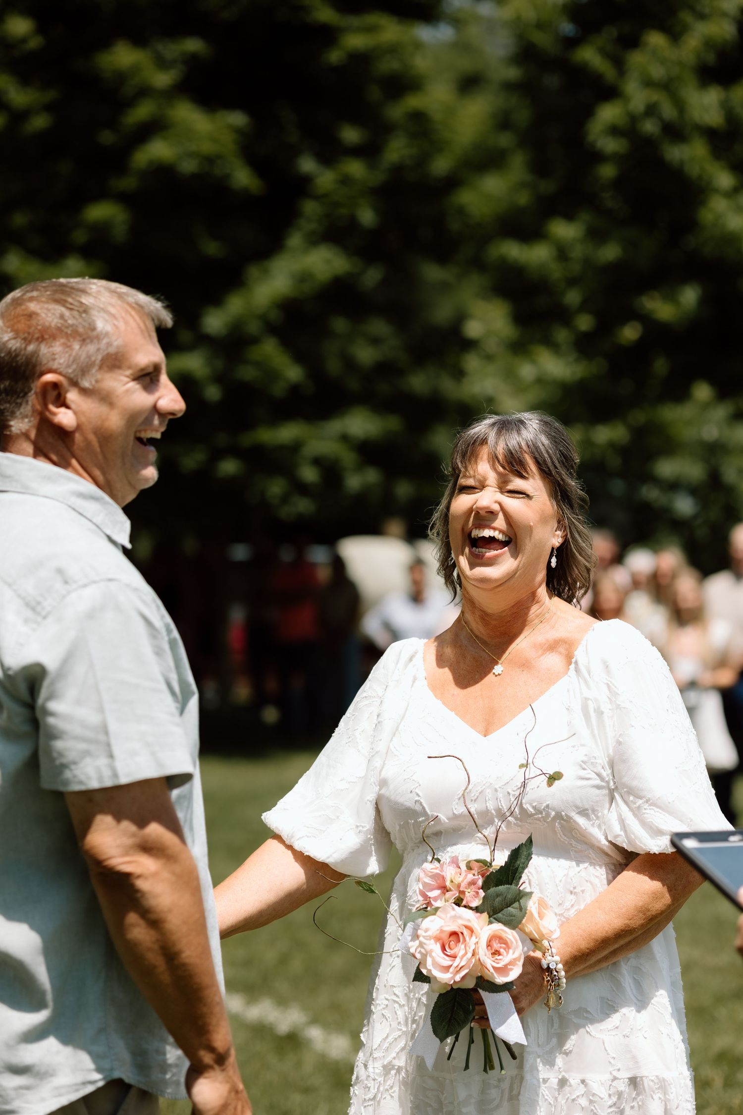 Laughing bride in white dress holds pink roses while standing with groom at outdoor wedding.