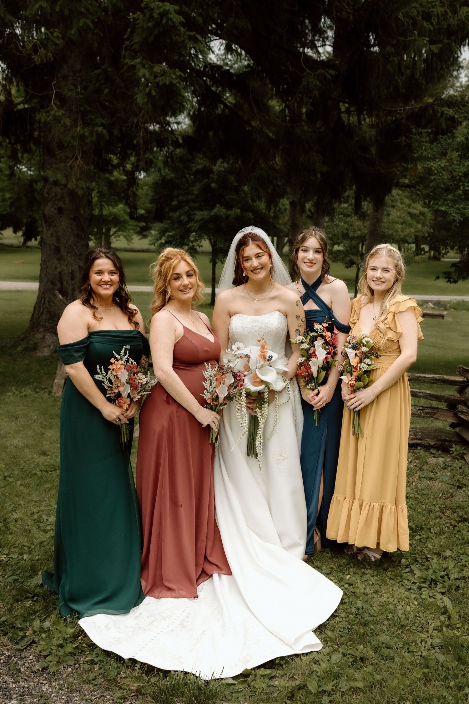 Bride in white gown poses with four bridesmaids in mismatched colorful dresses in park setting.