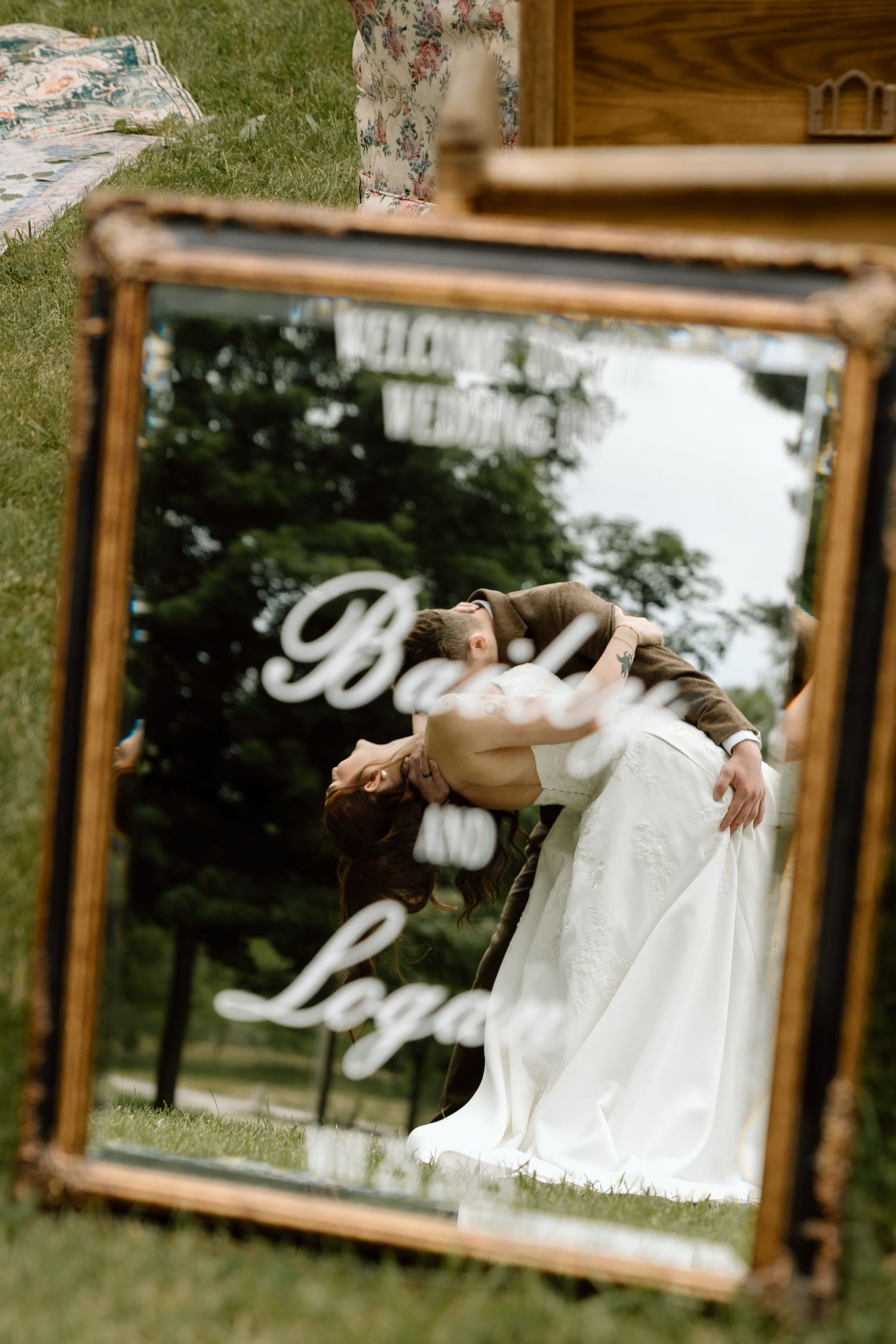 Bride and groom dipping reflected in ornate gold-framed wedding welcome mirror outdoors.