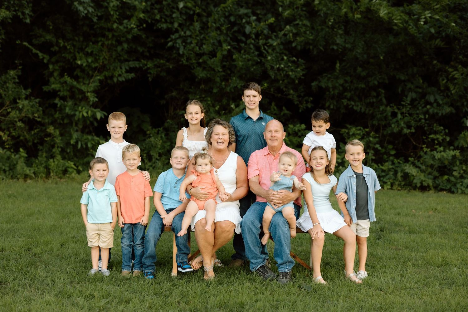 Grandparents pose outdoors on grass with their grandchildren for a large family portrait surrounded by lush green trees.