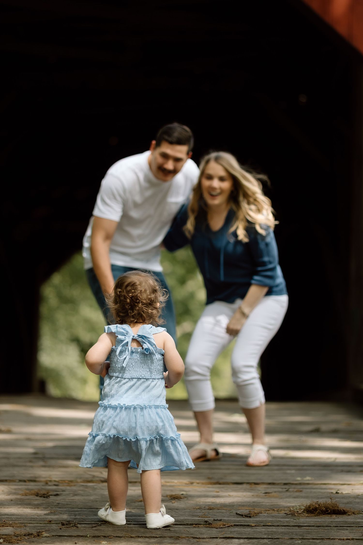 Little girl in blue dress runs toward her laughing parents on a rustic covered bridge during family photos.