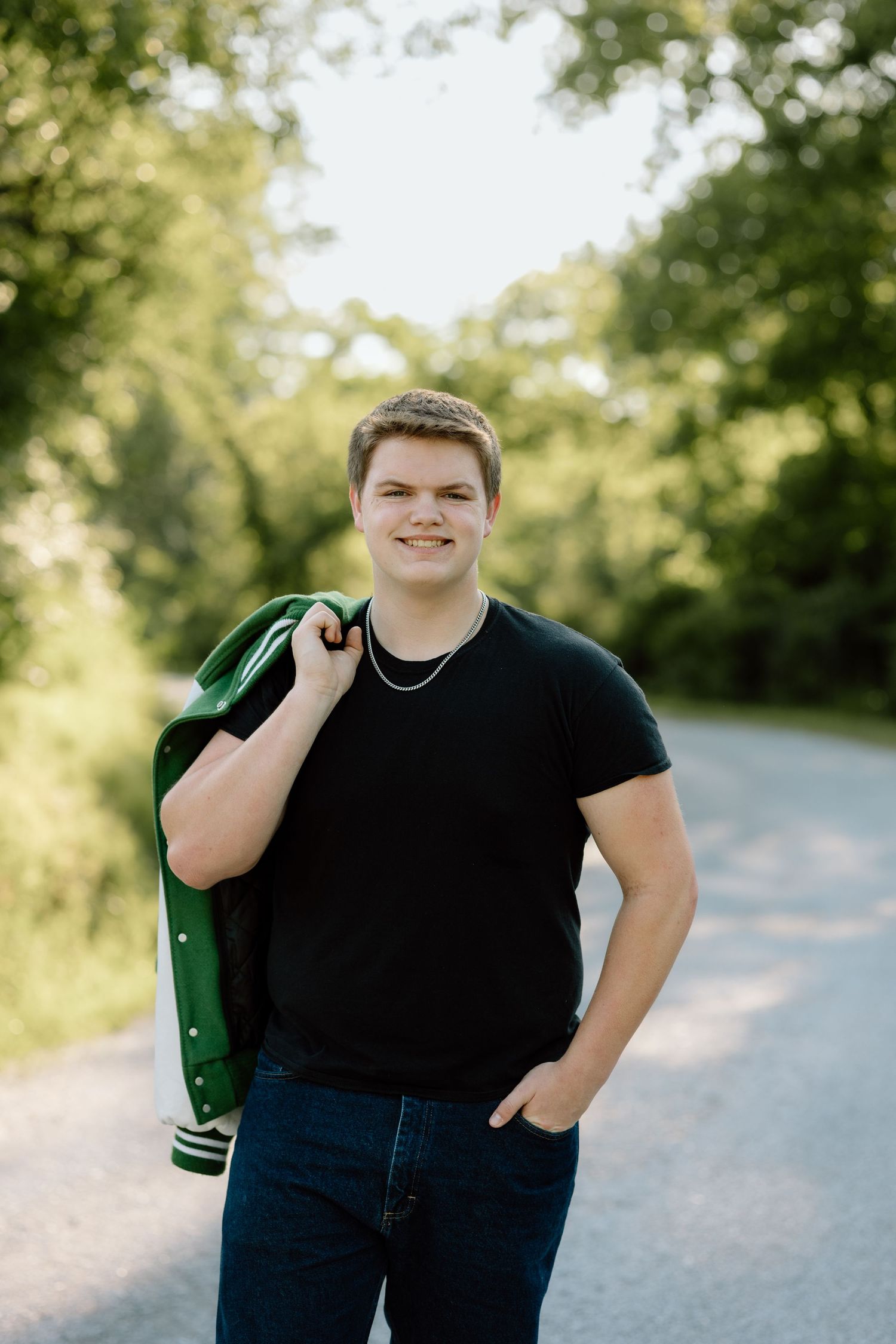 Smiling young man in black tee carries green letterman jacket on tree-lined country road.