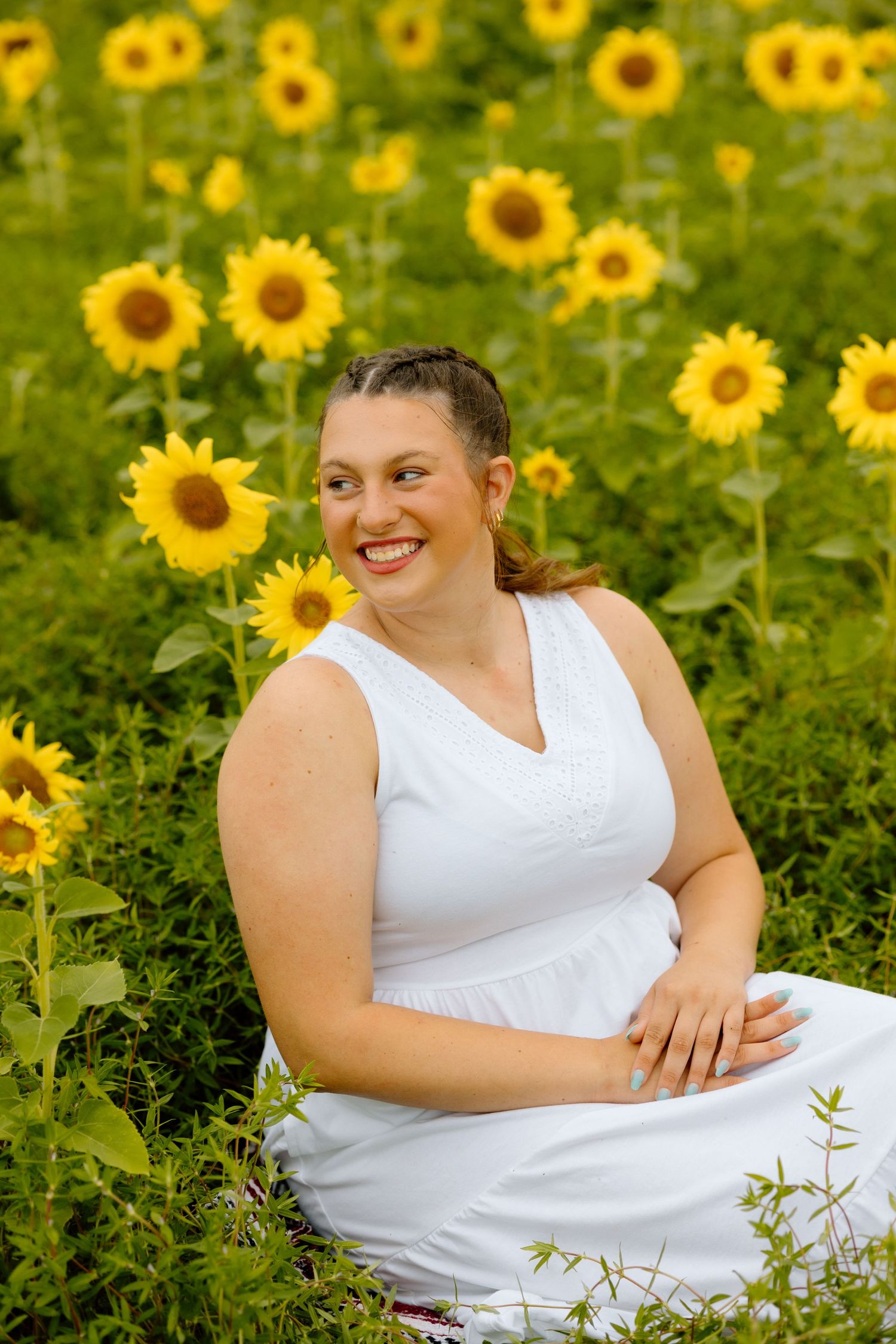 Smiling woman in white dress sitting among tall sunflowers in a lush green field.