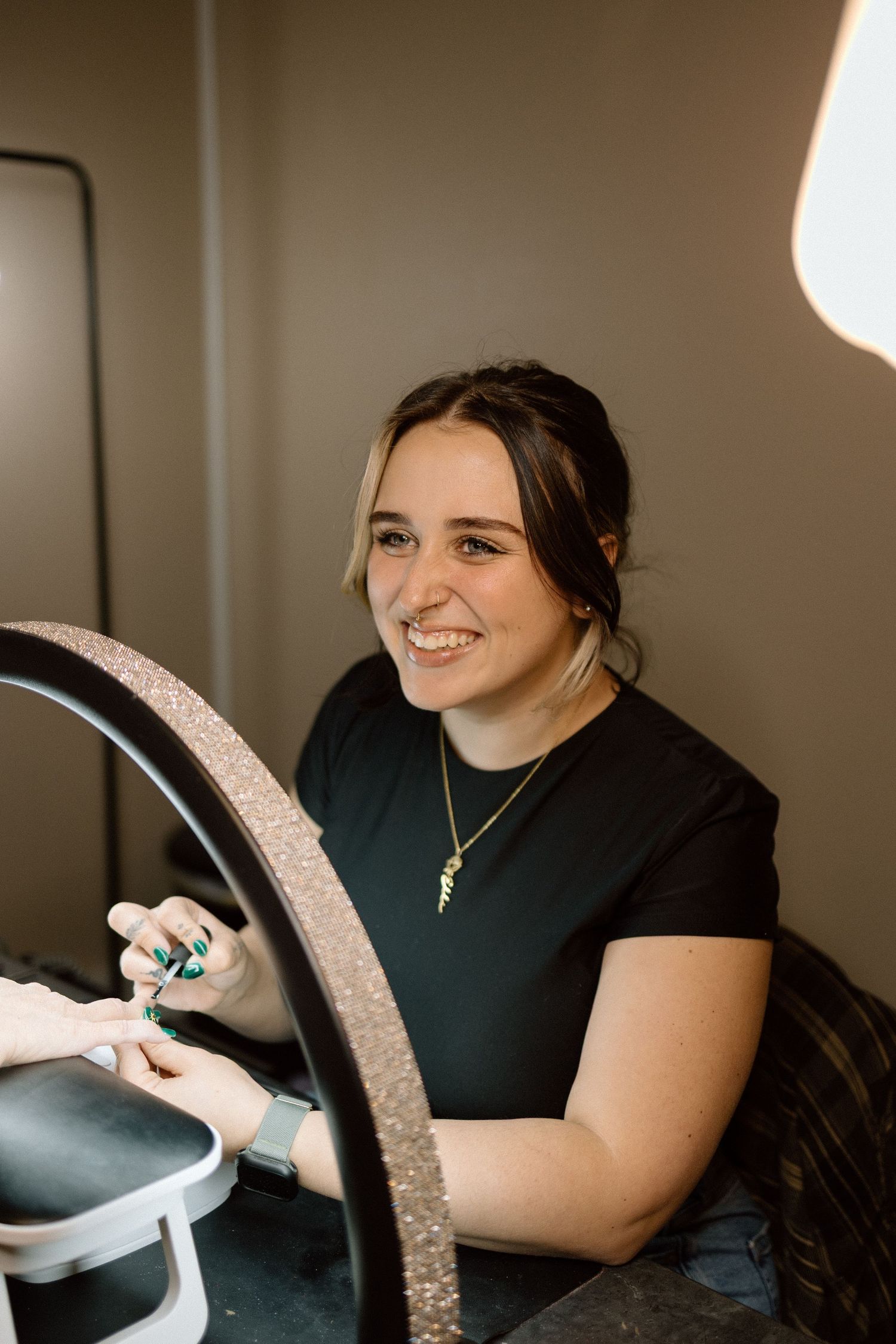 A smiling nail technician works on a client's nails under a ring light in a salon.