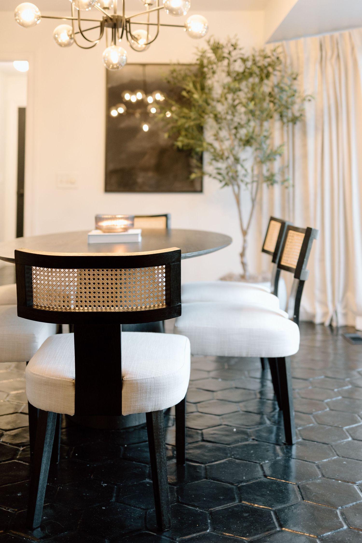 Modern dining room with round black table, cane-back chairs, white upholstery, chandelier, and olive tree.