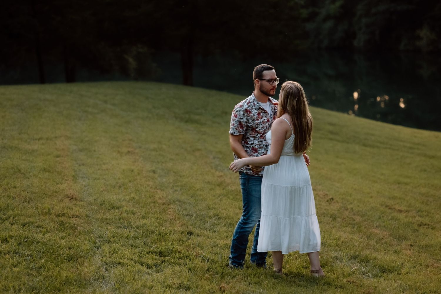 A couple stands close together in a grassy field at dusk, the woman in a white dress and man in a floral shirt.
