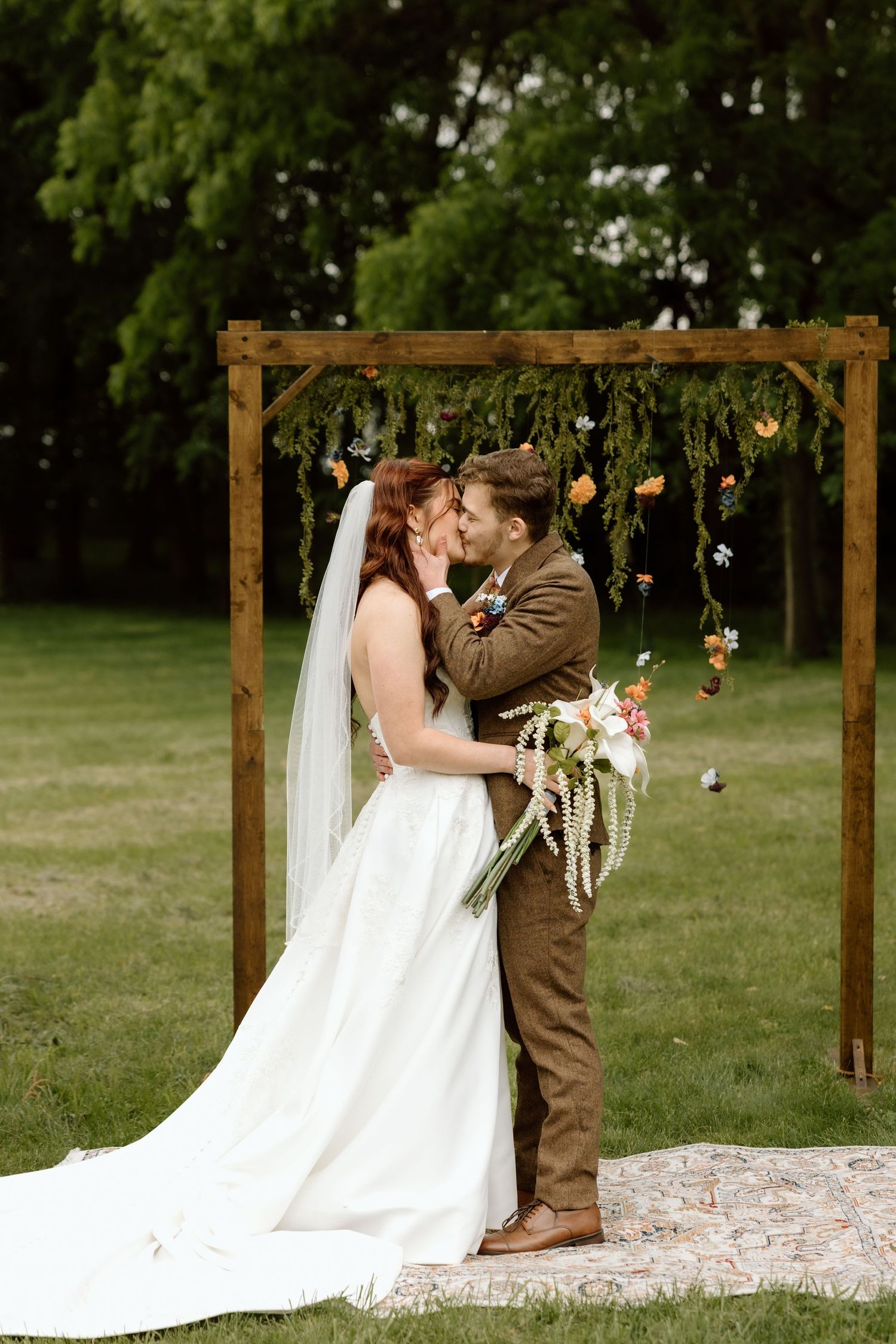 Bride and groom share first kiss under rustic wooden arch adorned with hanging greenery and colorful flowers.
