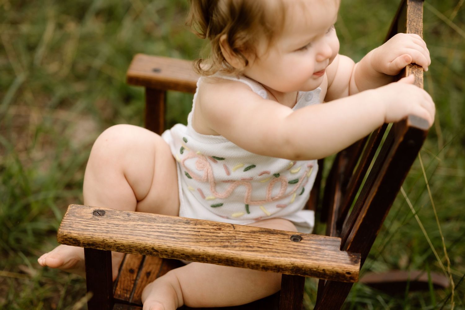 Baby girl in white floral romper climbing on small wooden chair outdoors in green grass.