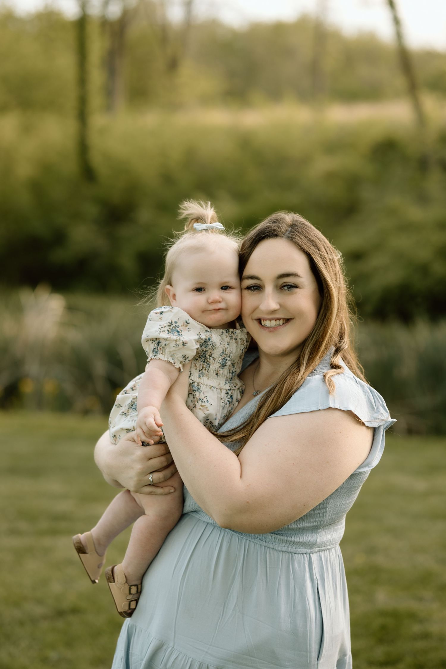 A smiling mother holds her happy baby girl close while posing outdoors in a beautiful green park setting.