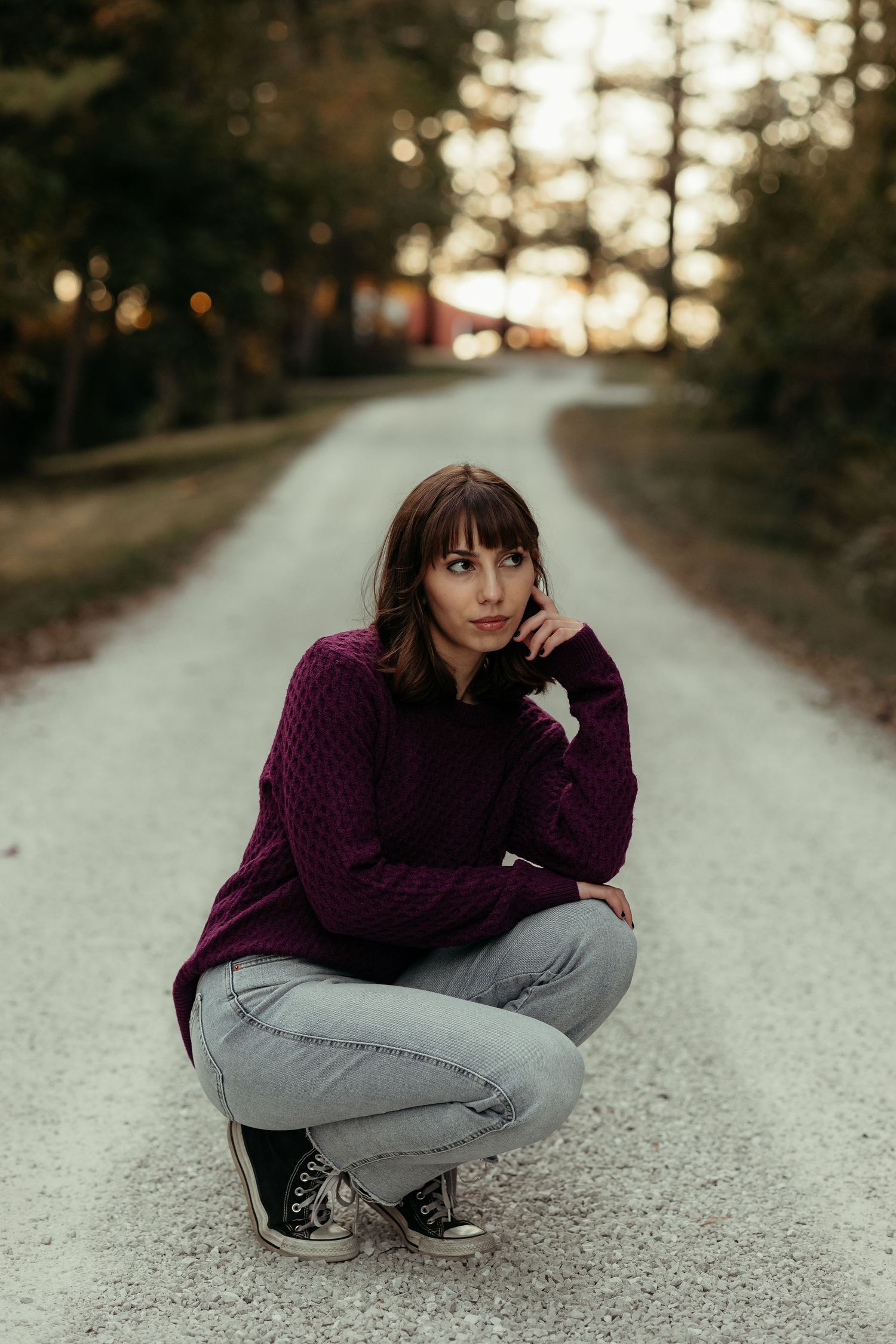 Young woman in purple sweater crouches on a quiet gravel road surrounded by autumn trees.