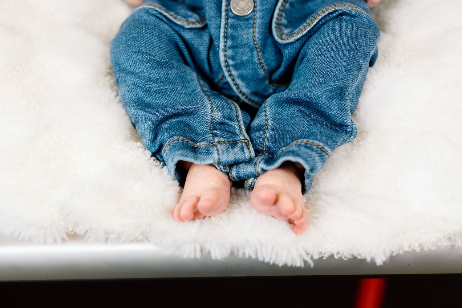 Newborn baby in denim outfit lying on a white fluffy blanket with tiny bare feet visible.