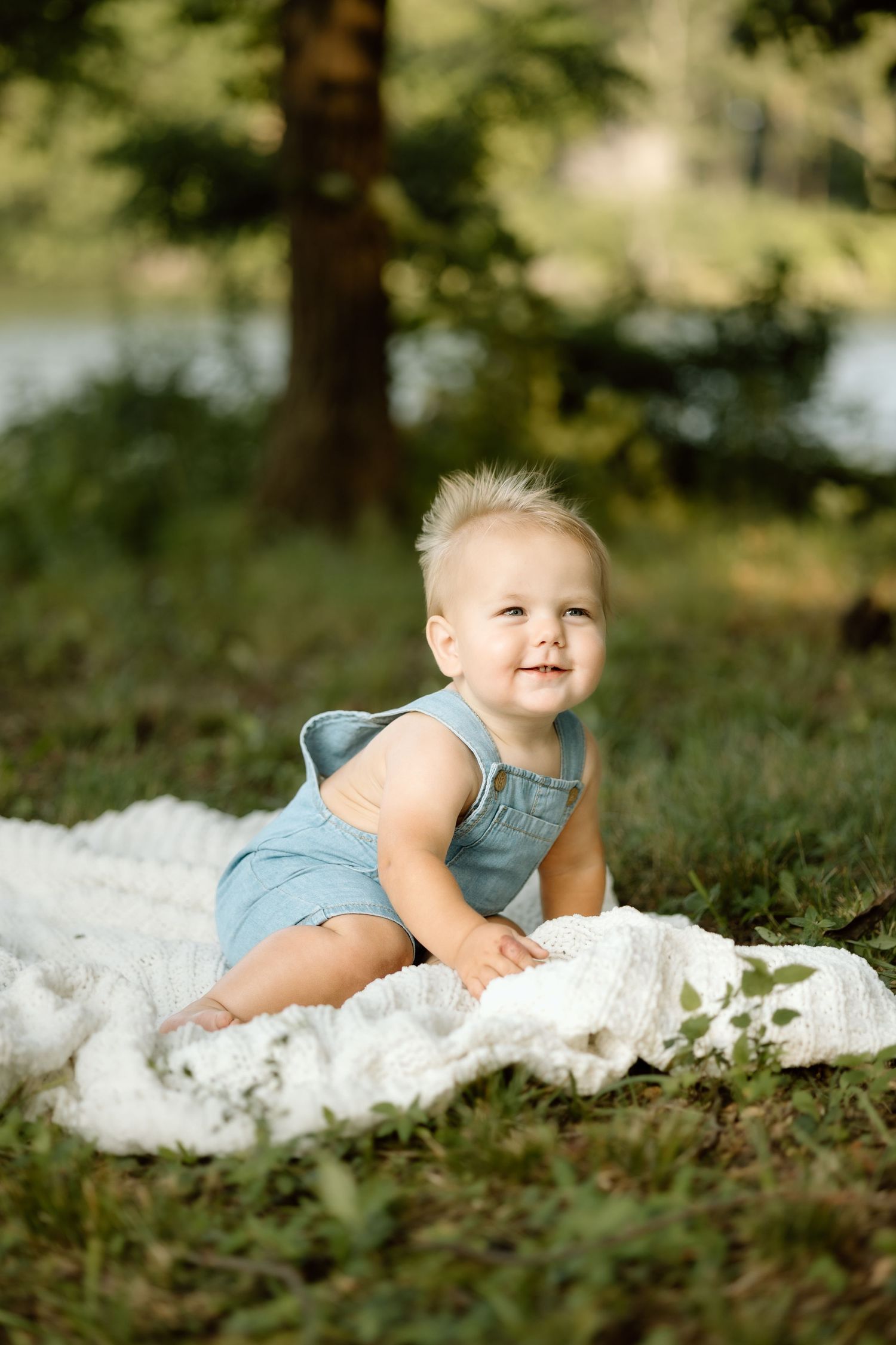 Smiling baby in blue romper sitting on white blanket in lush green park.