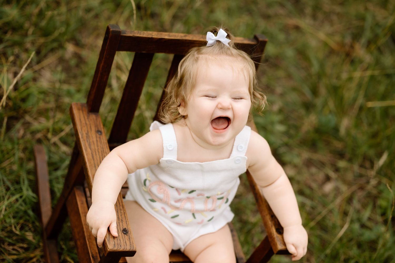 Laughing baby girl in white romper sitting on wooden chair outdoors in green grass.