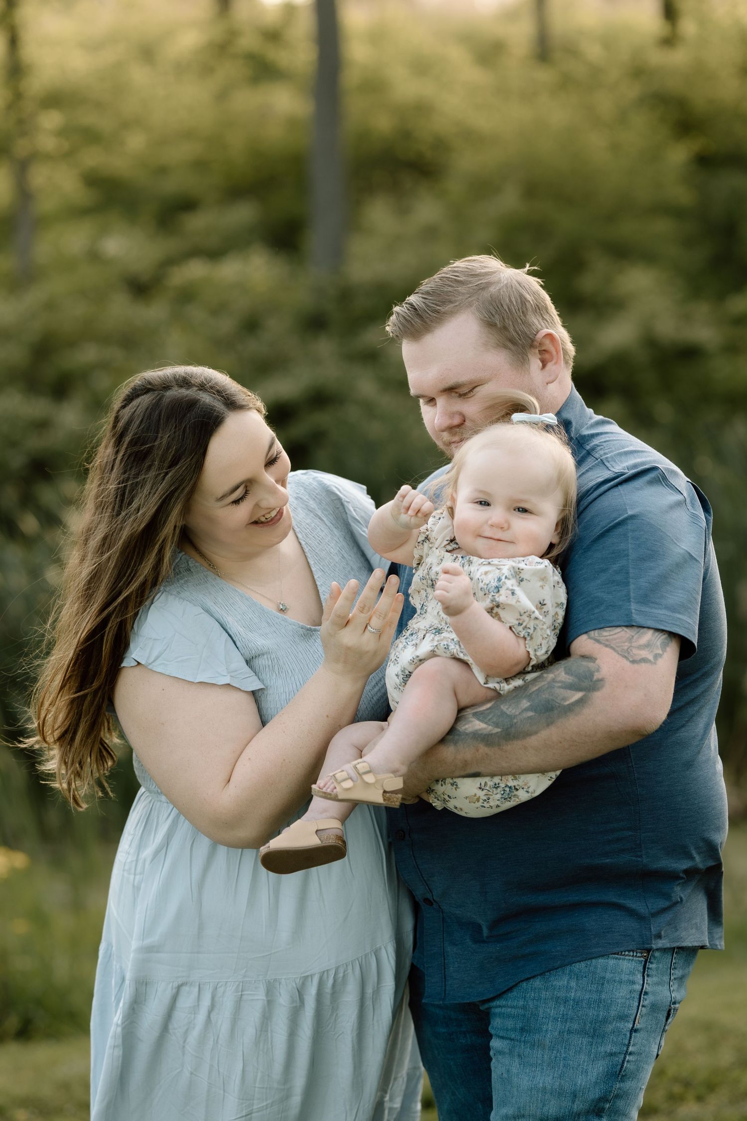 Loving parents interact playfully with their cheerful baby girl outdoors in a lush green garden setting.