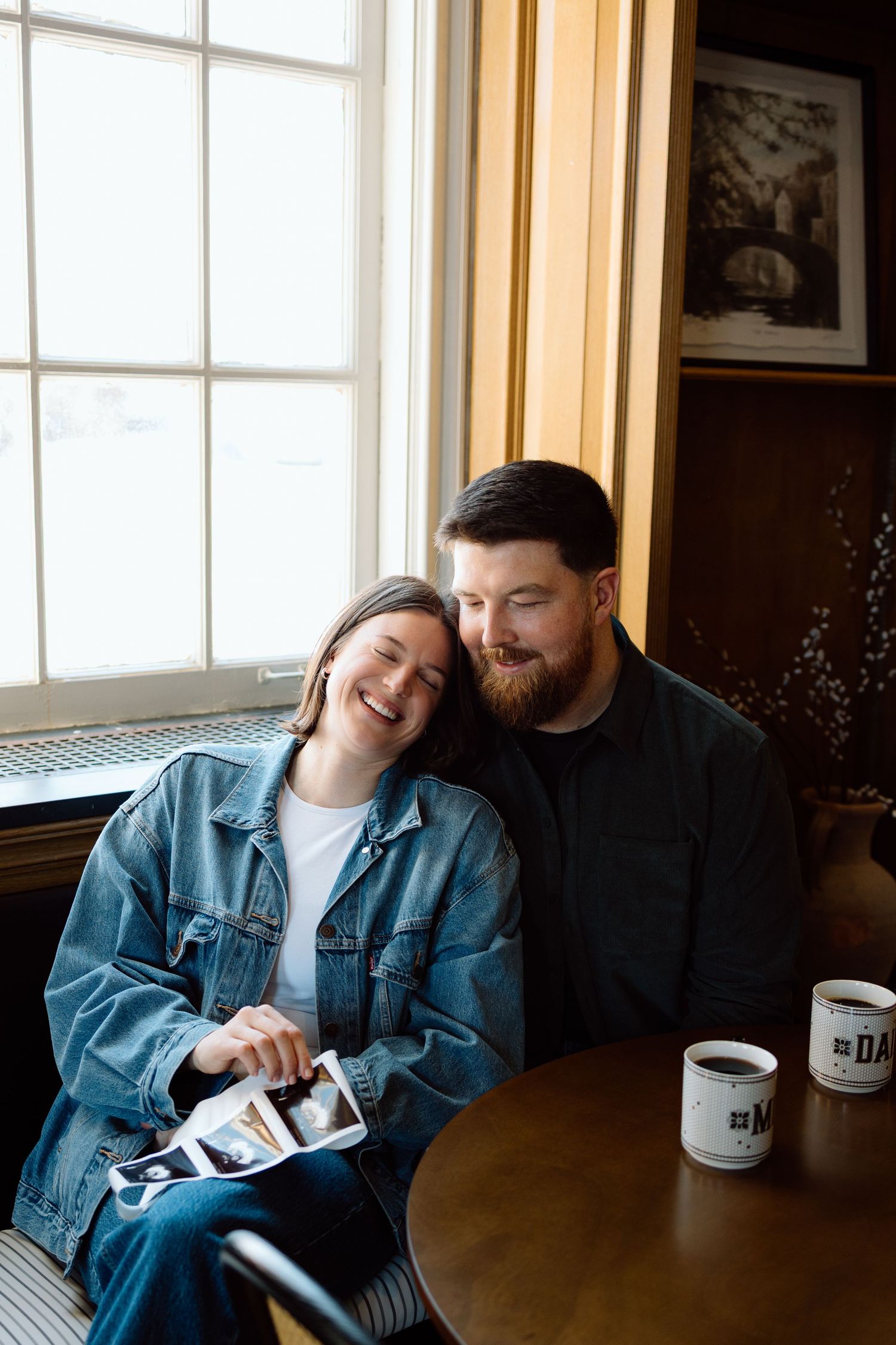 Happy couple snuggling at table with ultrasound photos and Mom and Dad mugs by window.