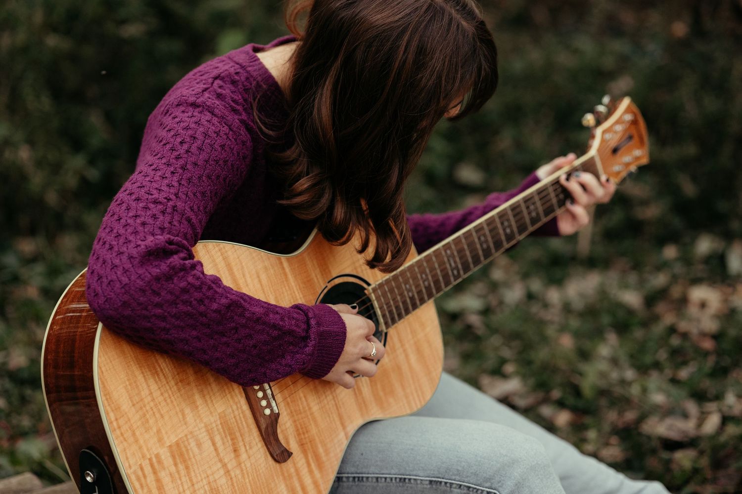 Young woman in purple sweater plays acoustic guitar outdoors in autumn forest.