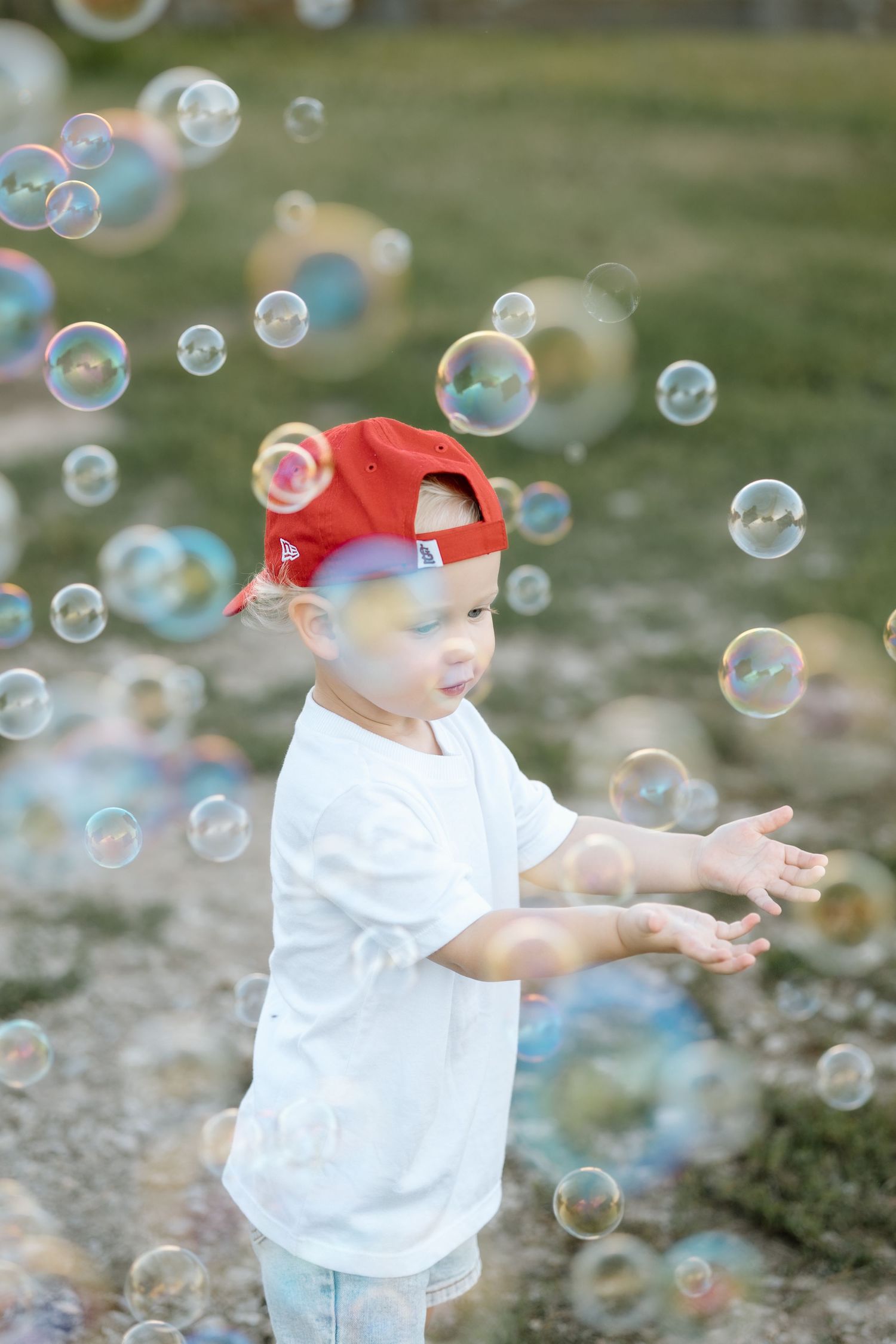 Happy toddler boy in red baseball cap reaching out to catch soap bubbles outside.