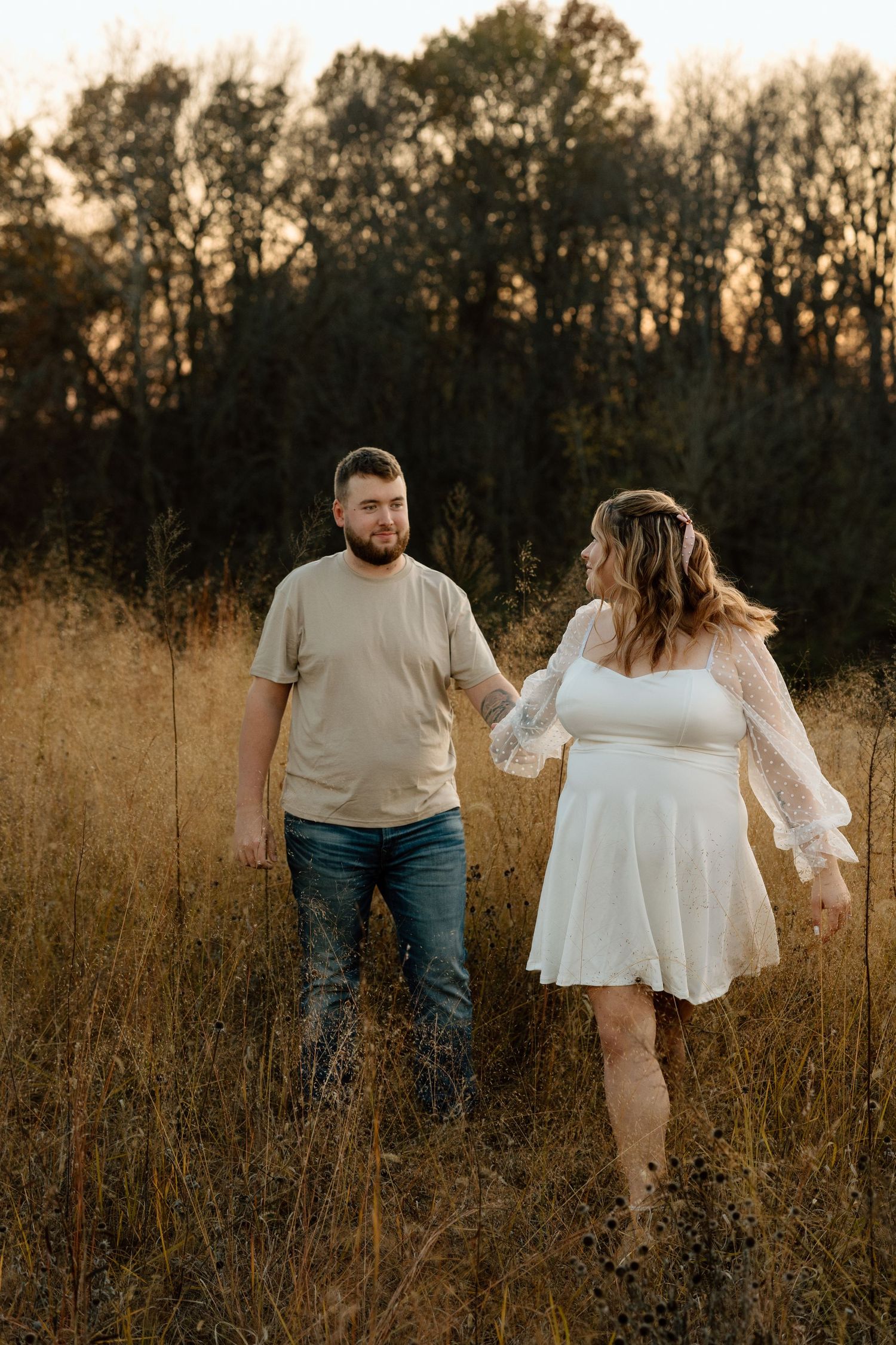 Couple walking hand in hand through tall golden grass field at dusk during fall engagement photo session.