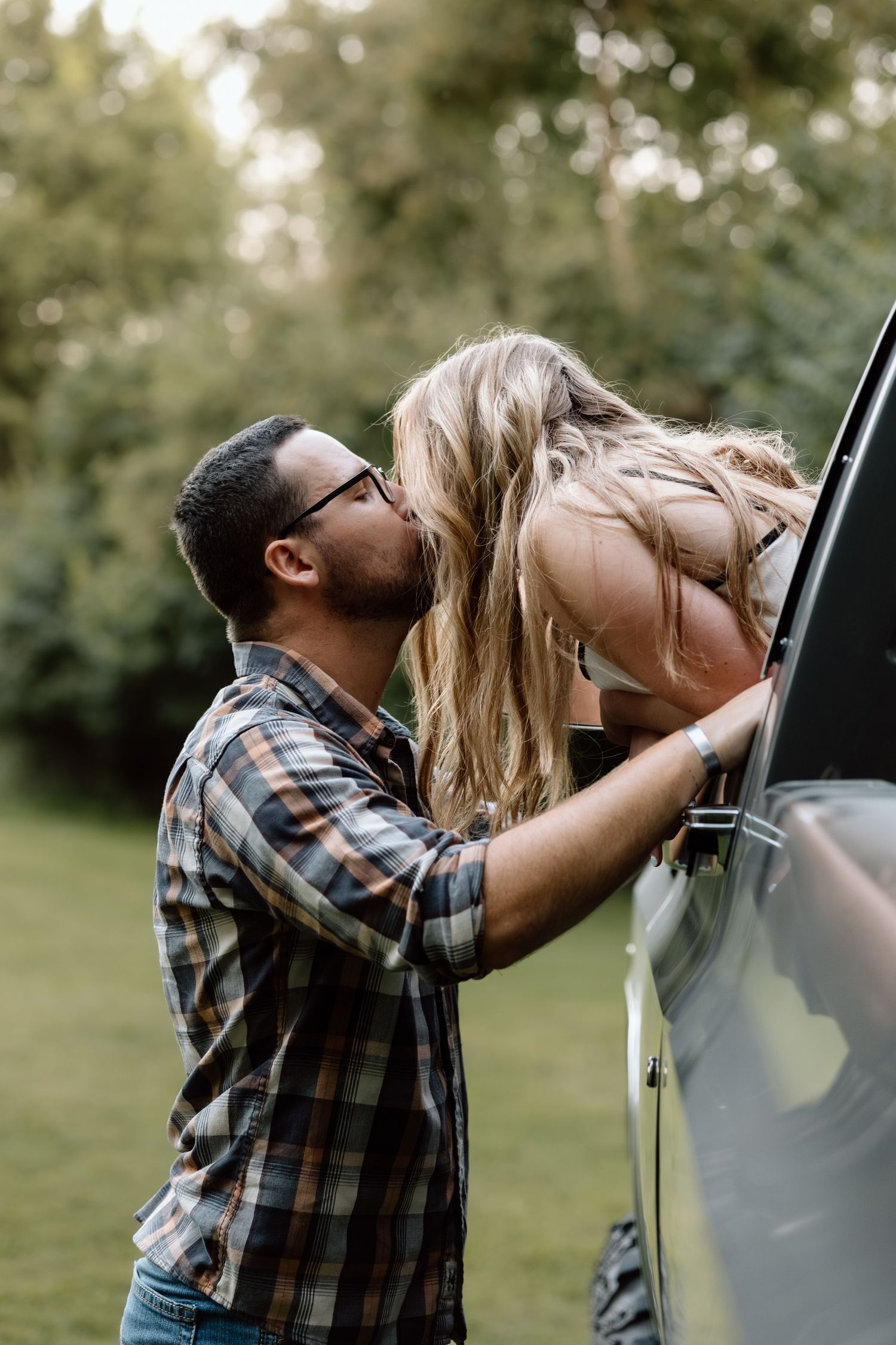 A man in a plaid shirt kisses a blonde woman leaning out of a car window in a lush green outdoor setting.