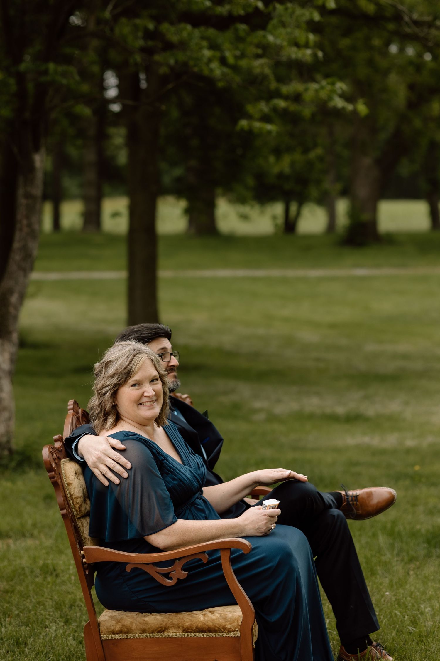 Couple poses outdoors in a lush green park, woman in teal dress seated in vintage chair with man behind her.