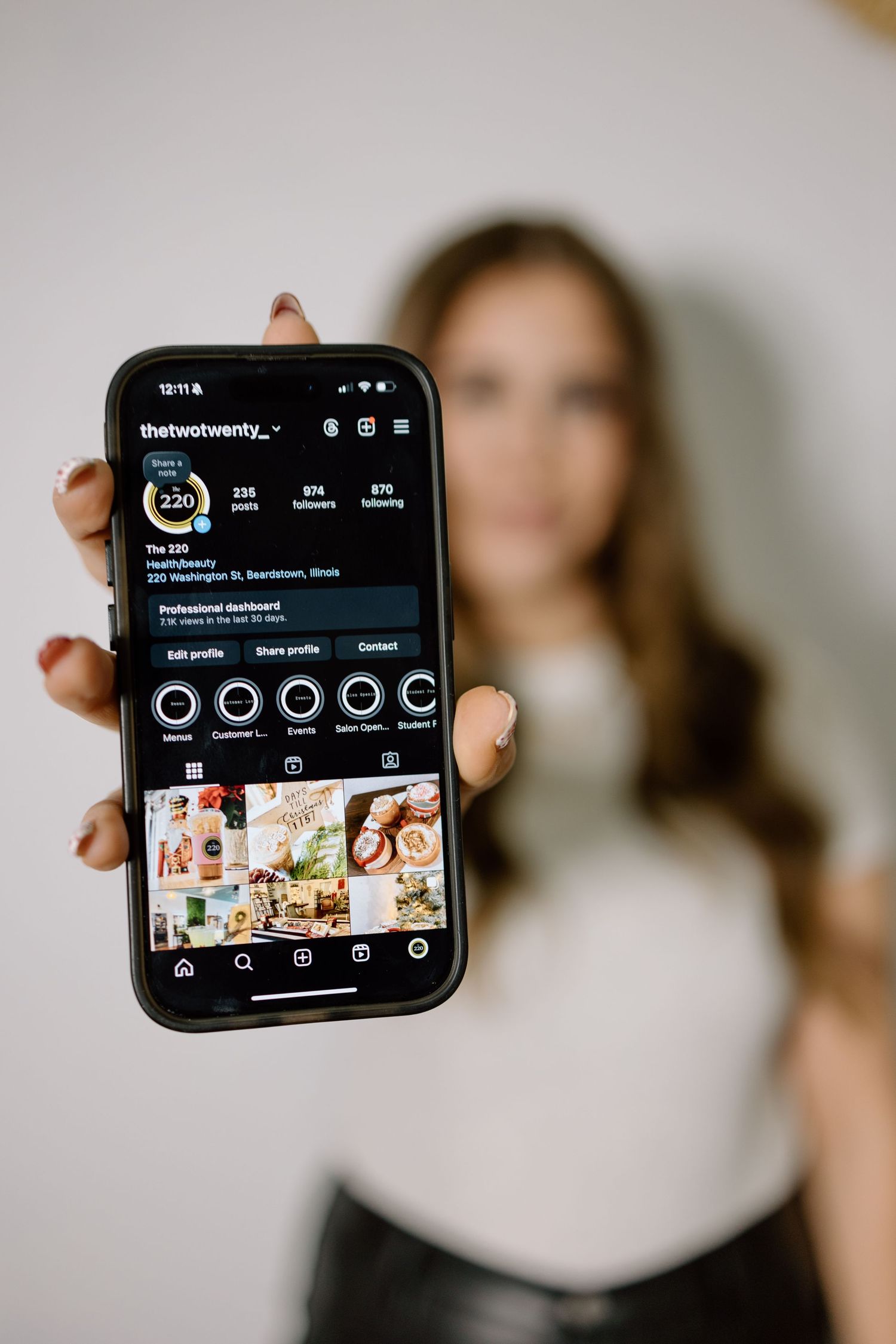 A woman holds up a smartphone displaying The 220 Salon Boutique Instagram profile page.