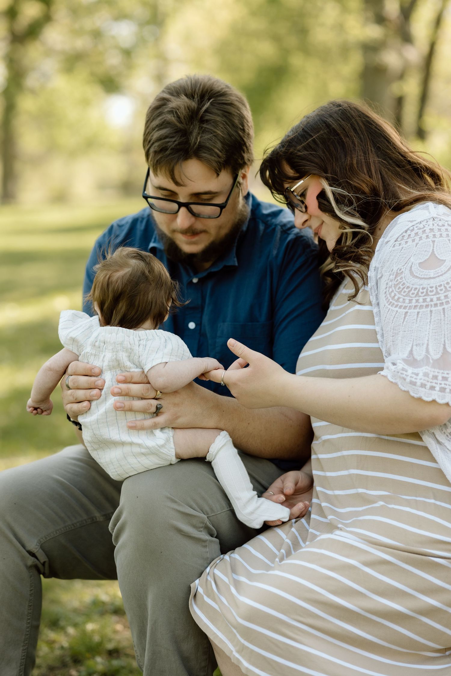 New parents gently hold their newborn baby while seated outdoors on a bench surrounded by lush greenery.