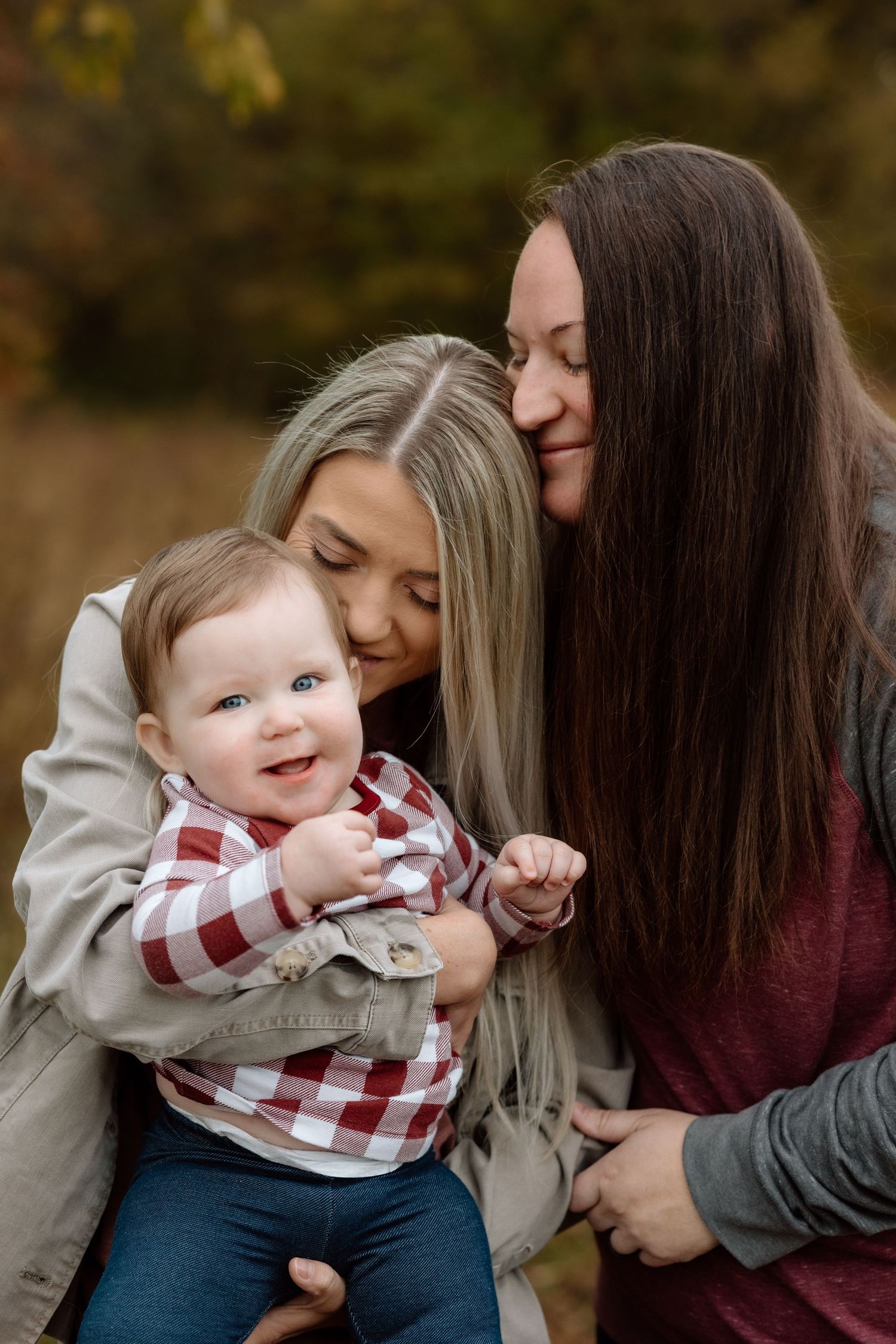 Two women tenderly kissing and cuddling a blue-eyed baby in a plaid shirt among autumn foliage.