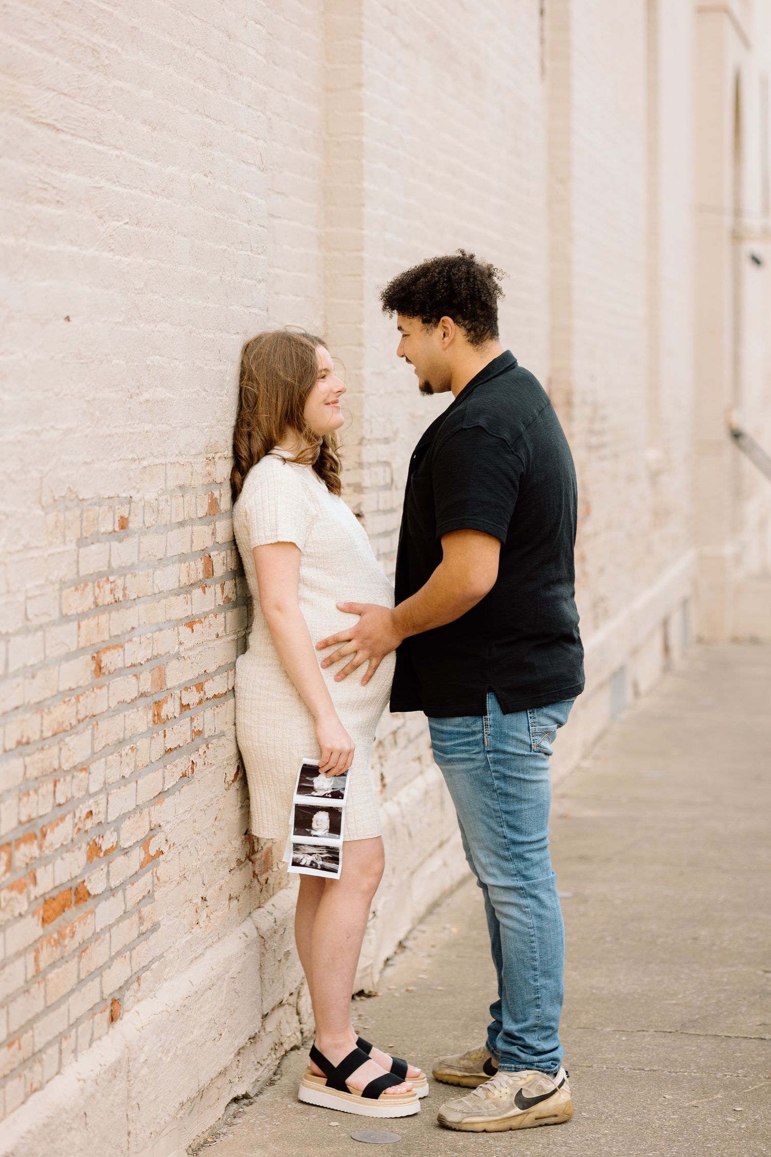 Expecting couple sharing a smile while holding ultrasound photos against brick wall during maternity session.