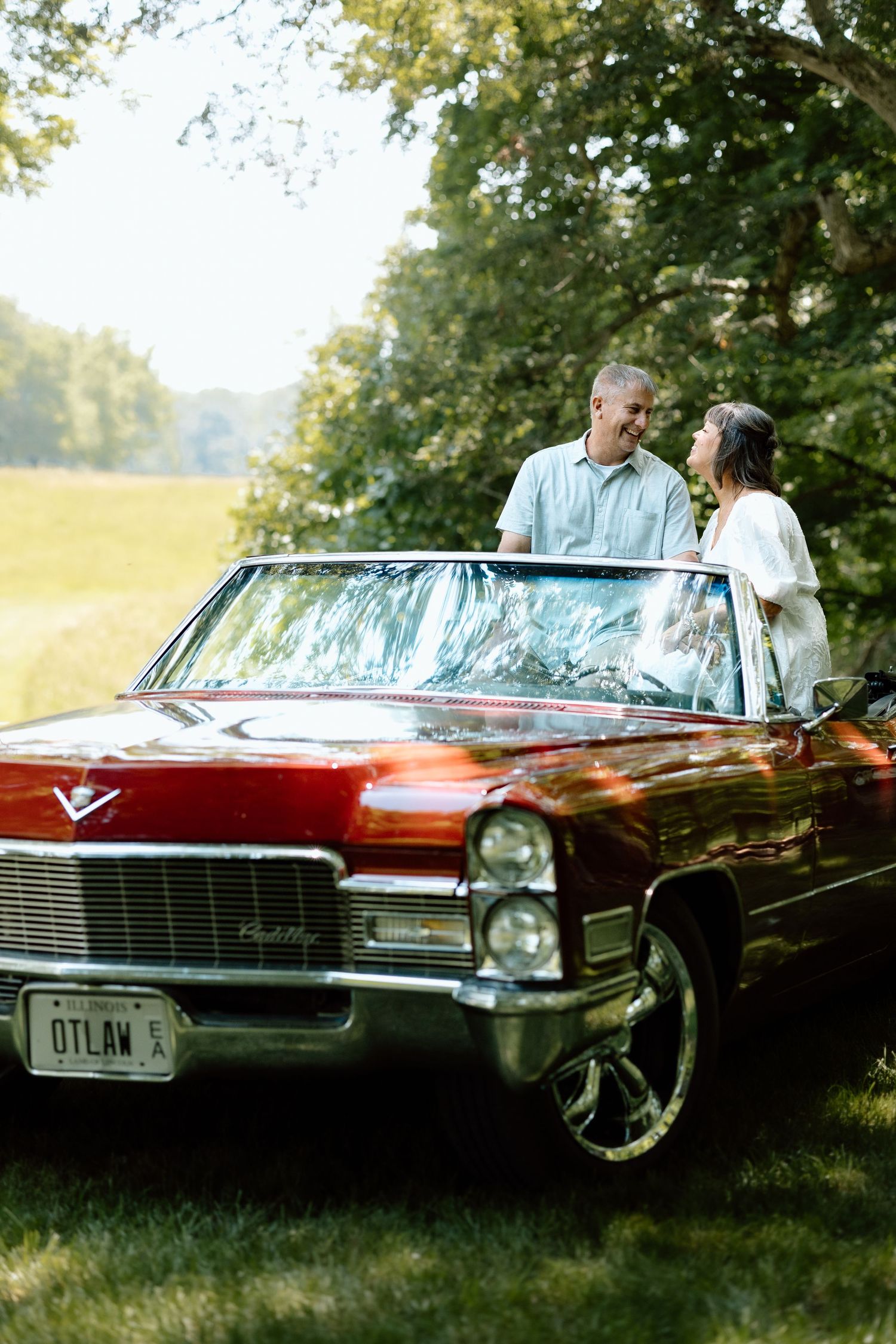 Couple smiling at each other standing beside classic red Cadillac convertible in sunny green field.