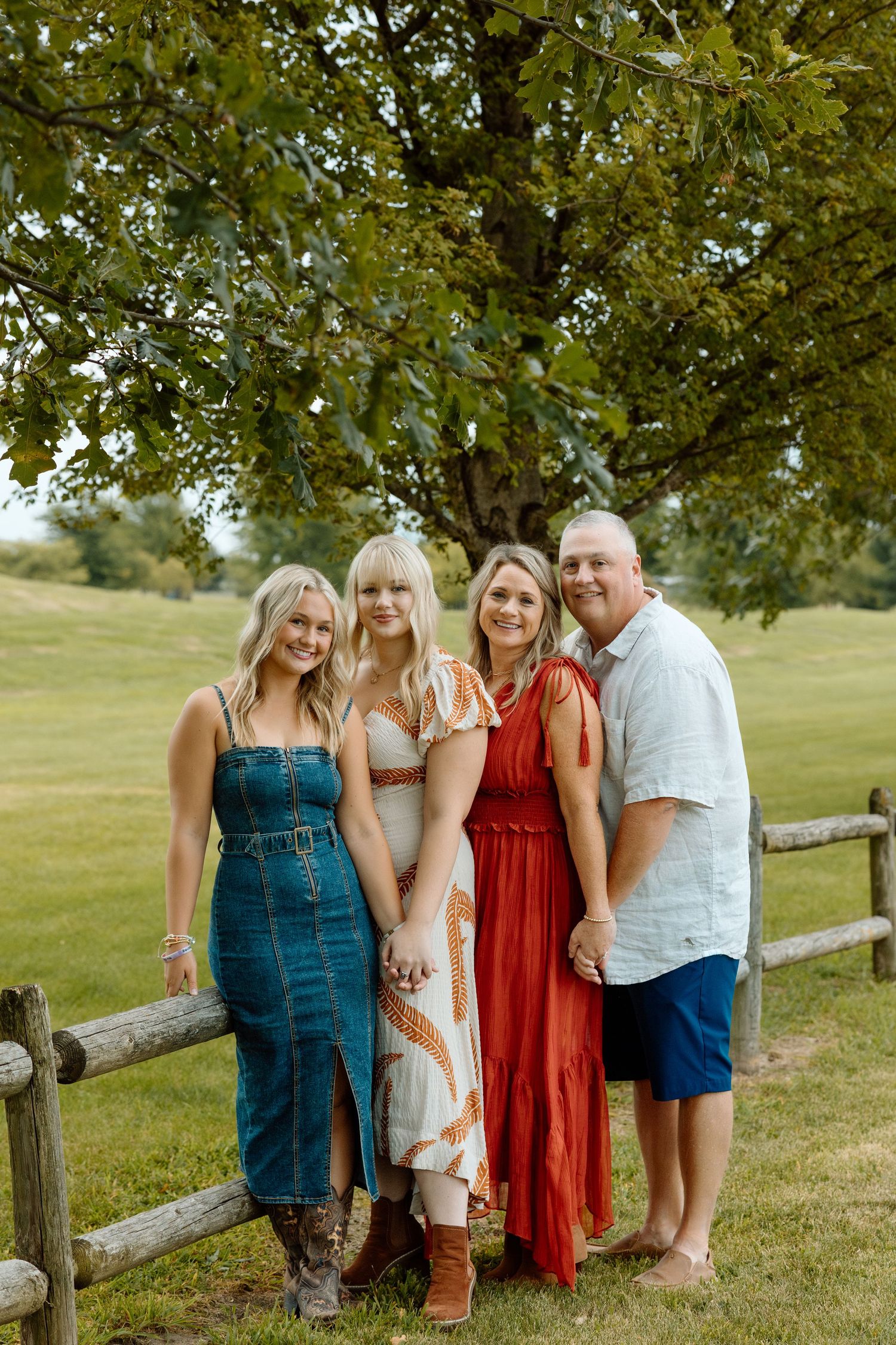 Family of four posing by a rustic wooden fence under a large tree in a green grassy park.