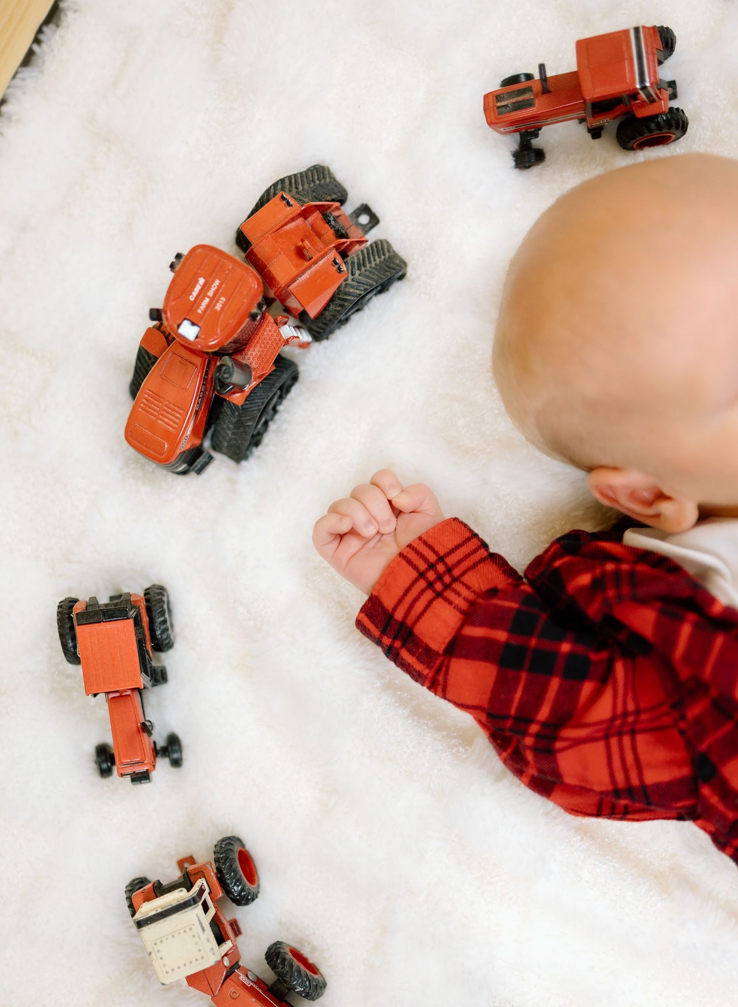 Overhead view of baby in red plaid flannel shirt sleeping on white fur rug surrounded by orange toy tractors.