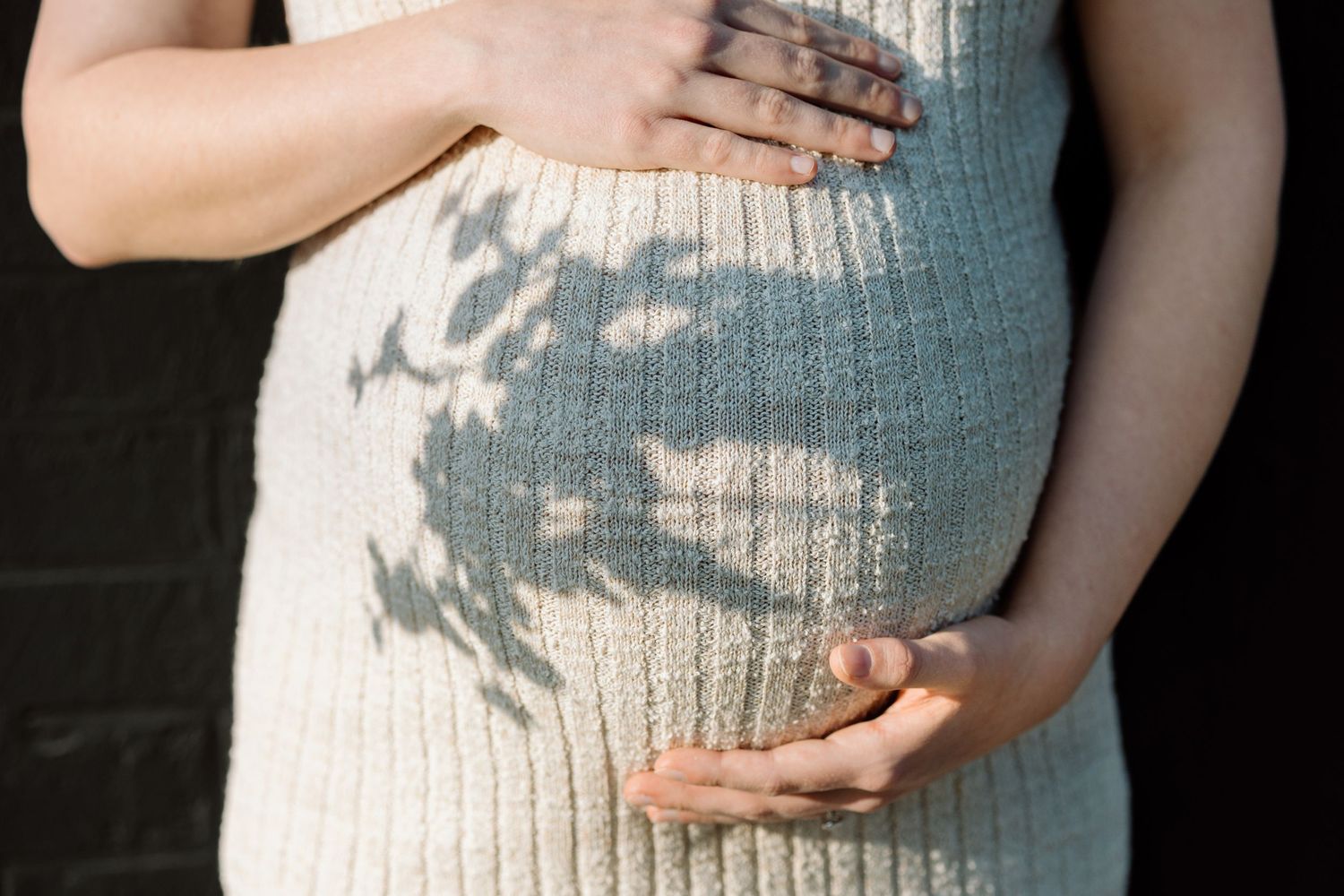 Close-up of pregnant belly in white knit dress with beautiful leaf shadow patterns in natural light.