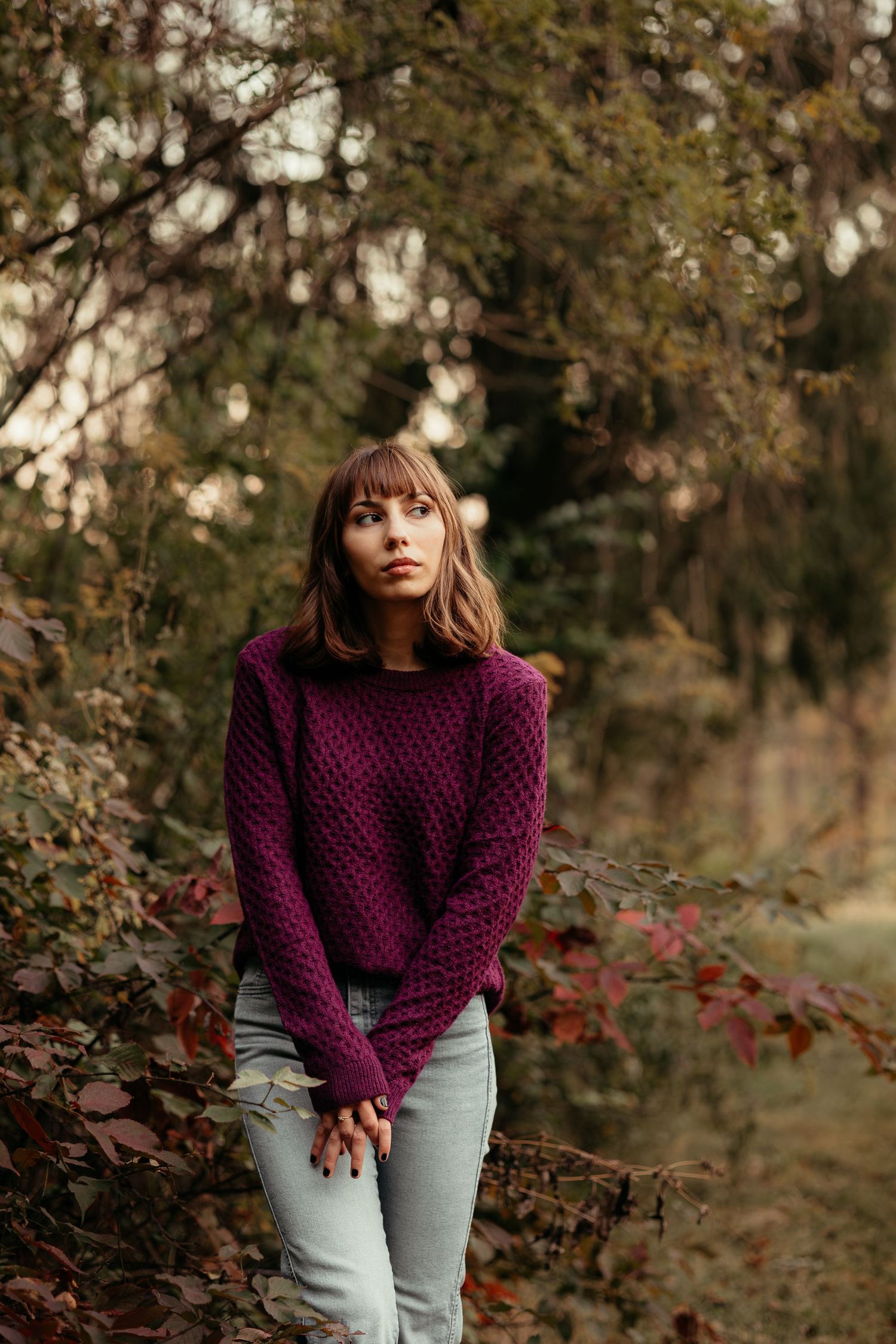 Young woman in purple cable-knit sweater standing among colorful autumn leaves in a forest setting.