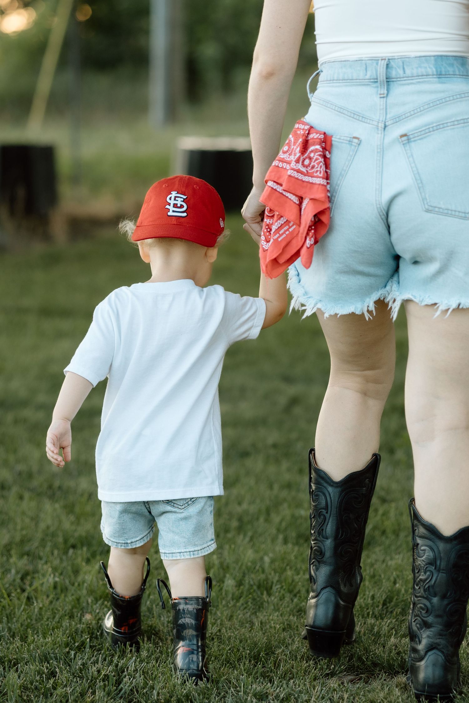 Toddler in St. Louis Cardinals cap holding mothers hand while walking on green grass.