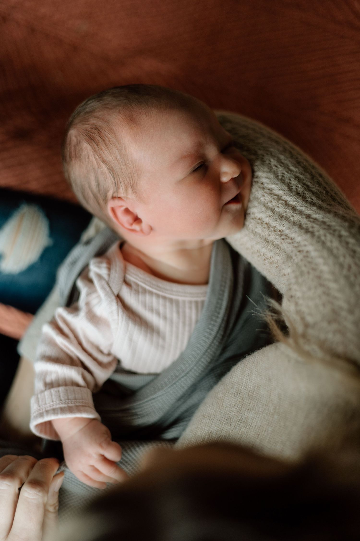 Awake newborn baby in white onesie and gray vest looking up while nestled in a cozy blanket.