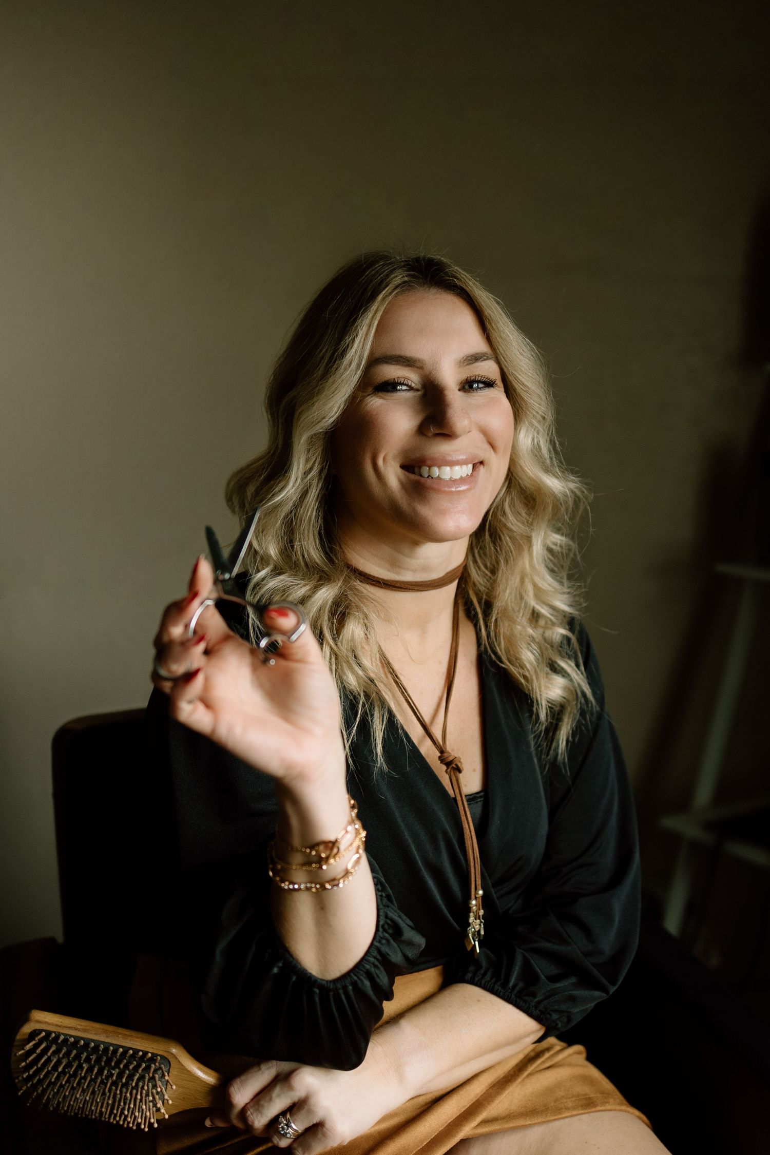 Smiling blonde hairstylist holding scissors while seated in a salon chair with warm lighting.