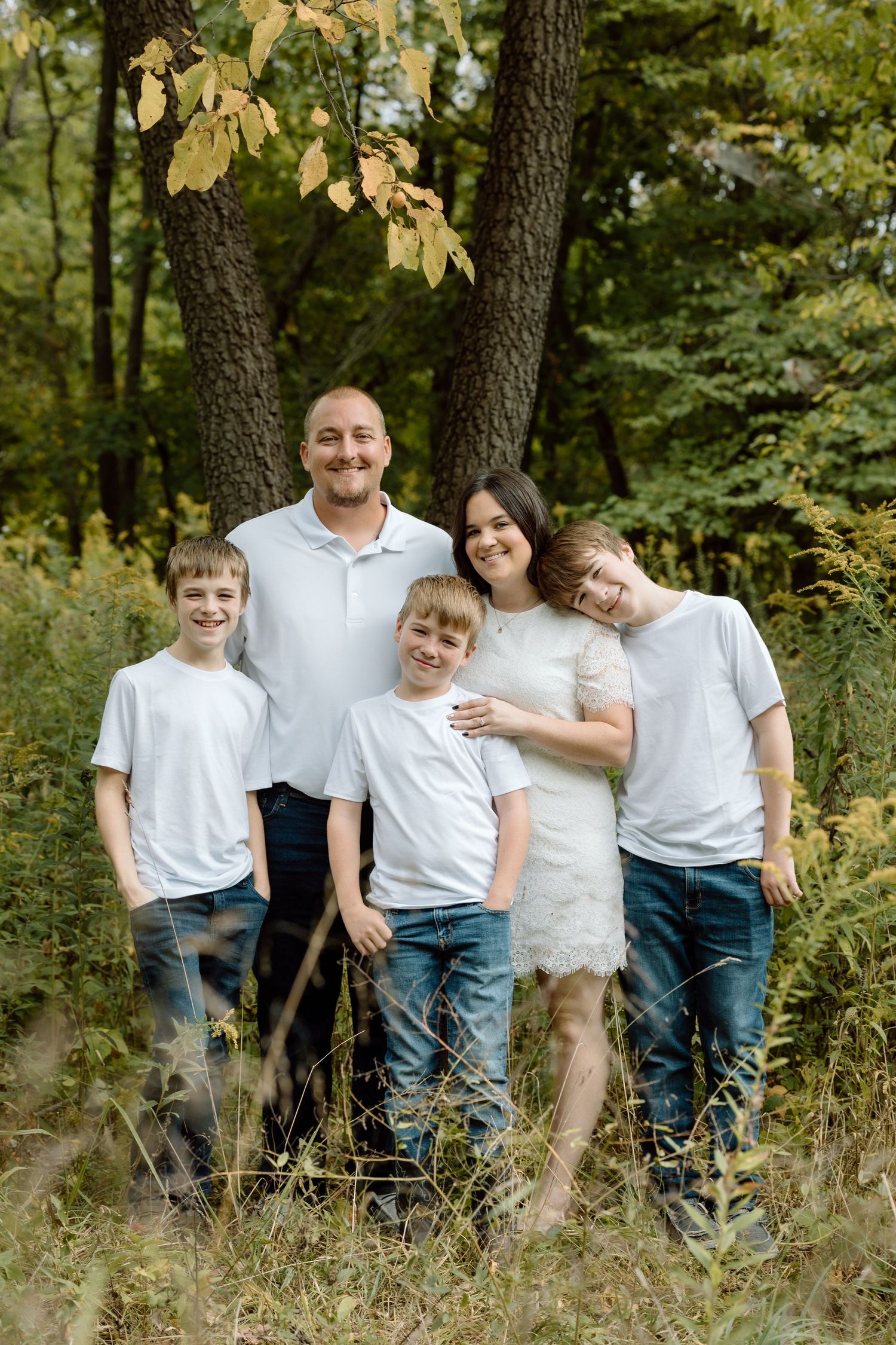 Happy family of five poses in white shirts and jeans among lush green trees in an outdoor forest setting.