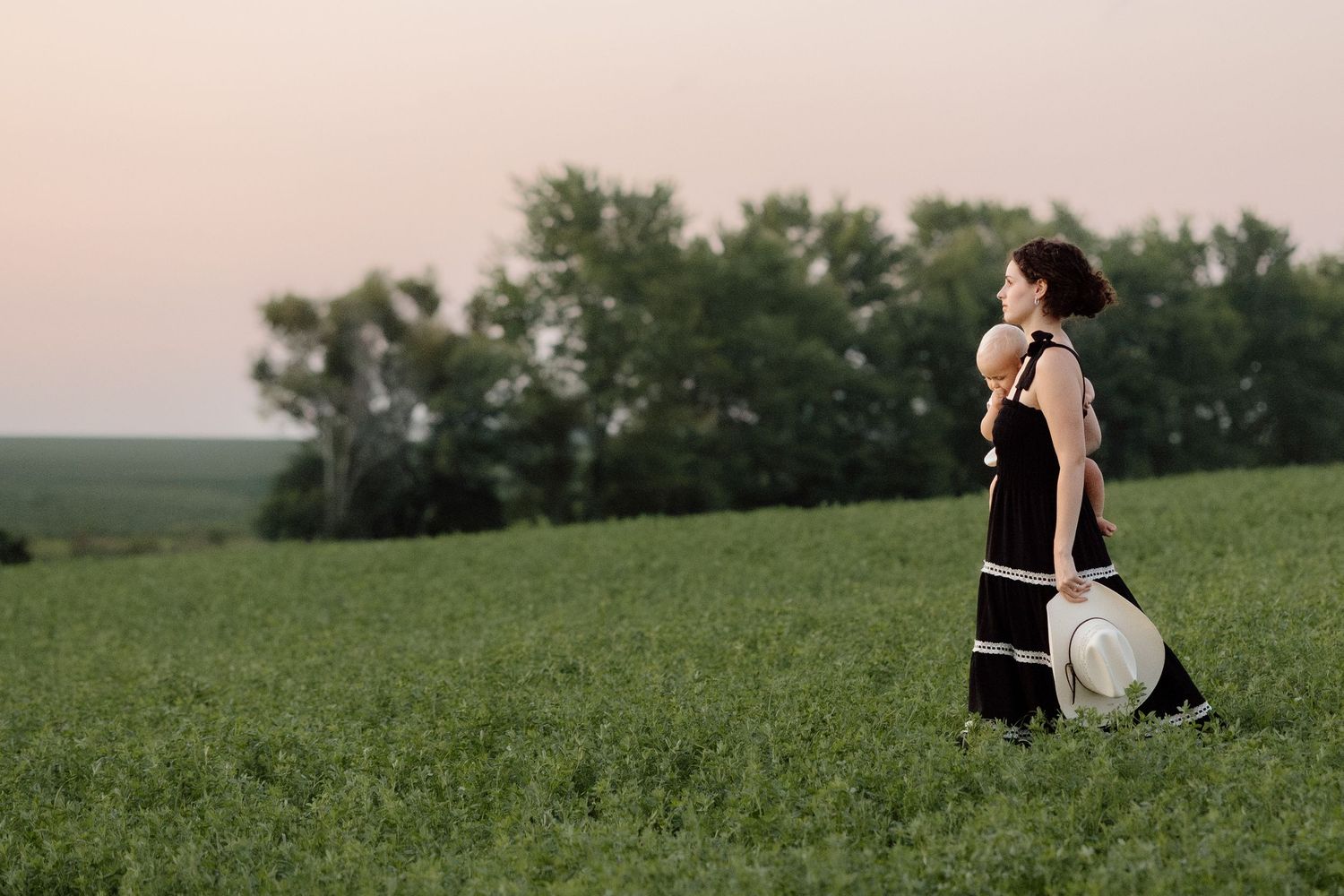 Woman in black dress holding a hat walking through a lush green field at sunset with trees in background.