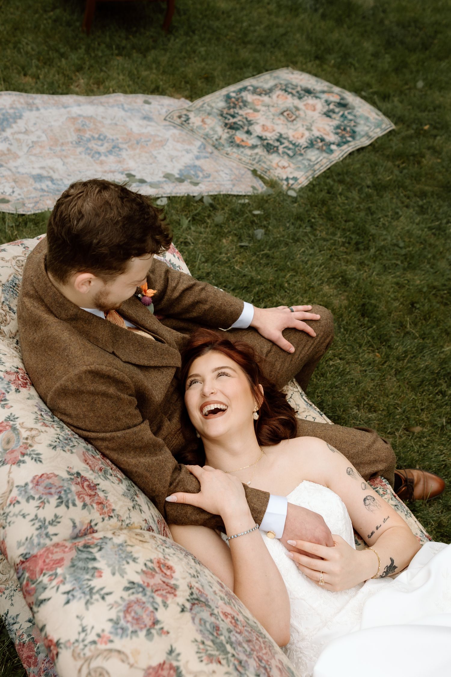 Laughing bride in white dress reclines on floral cushion with groom in brown suit outdoors.
