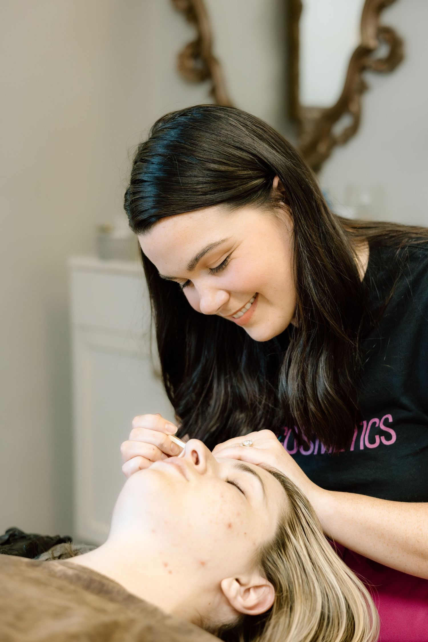 Smiling HR Cosmetics esthetician performing eyebrow grooming service on a blonde client in a beauty salon.