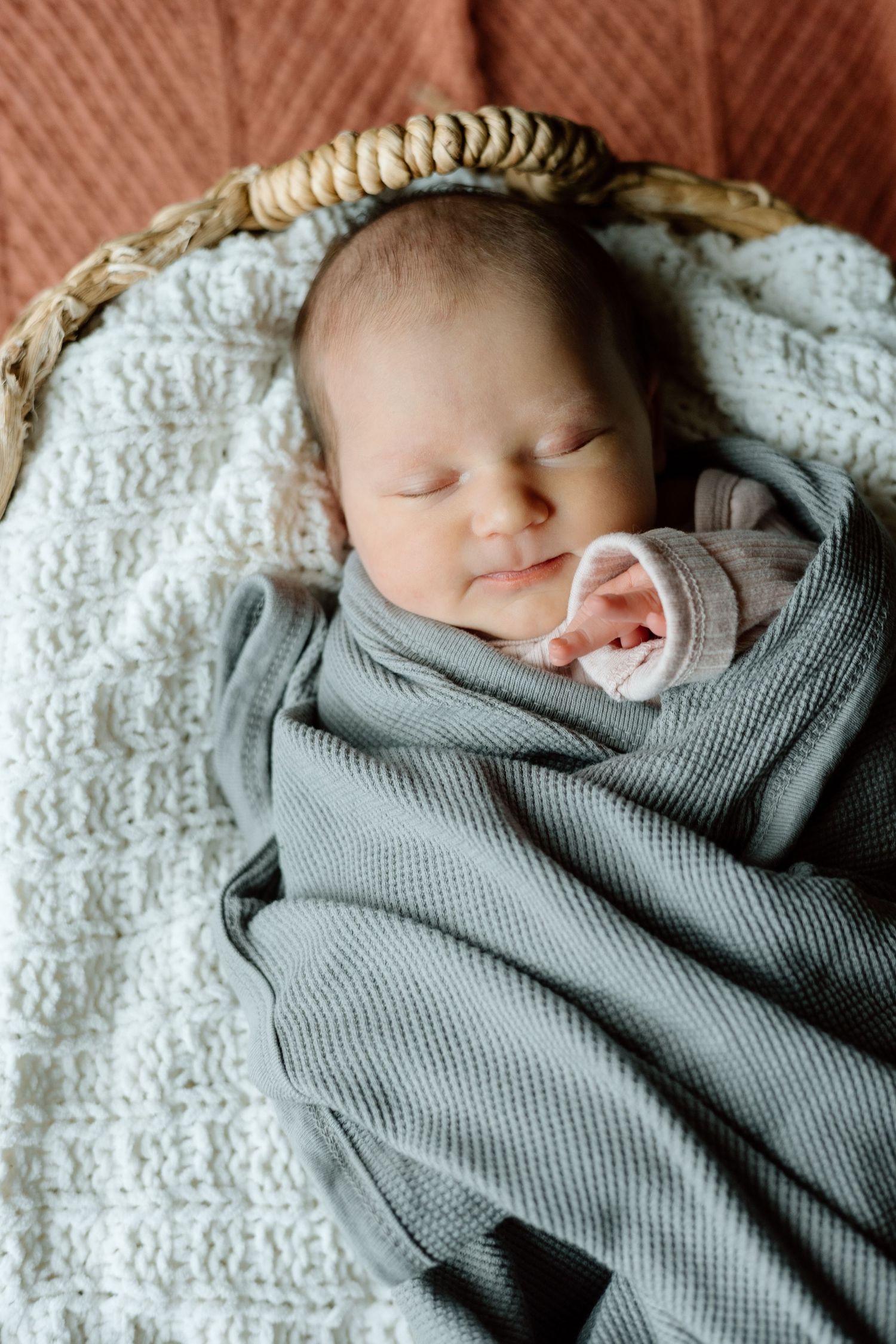 Sleeping newborn baby swaddled in gray blanket lying in a wicker basket with white knit lining.