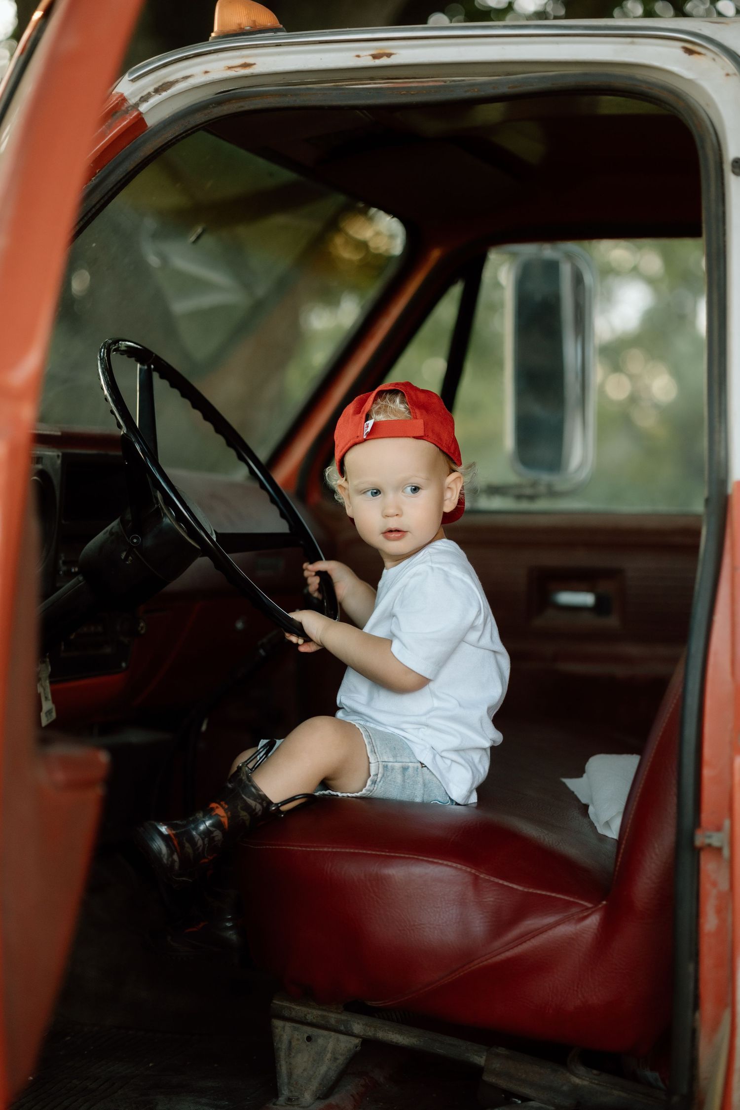 Toddler boy in red cap sitting behind the steering wheel of a vintage red farm truck.