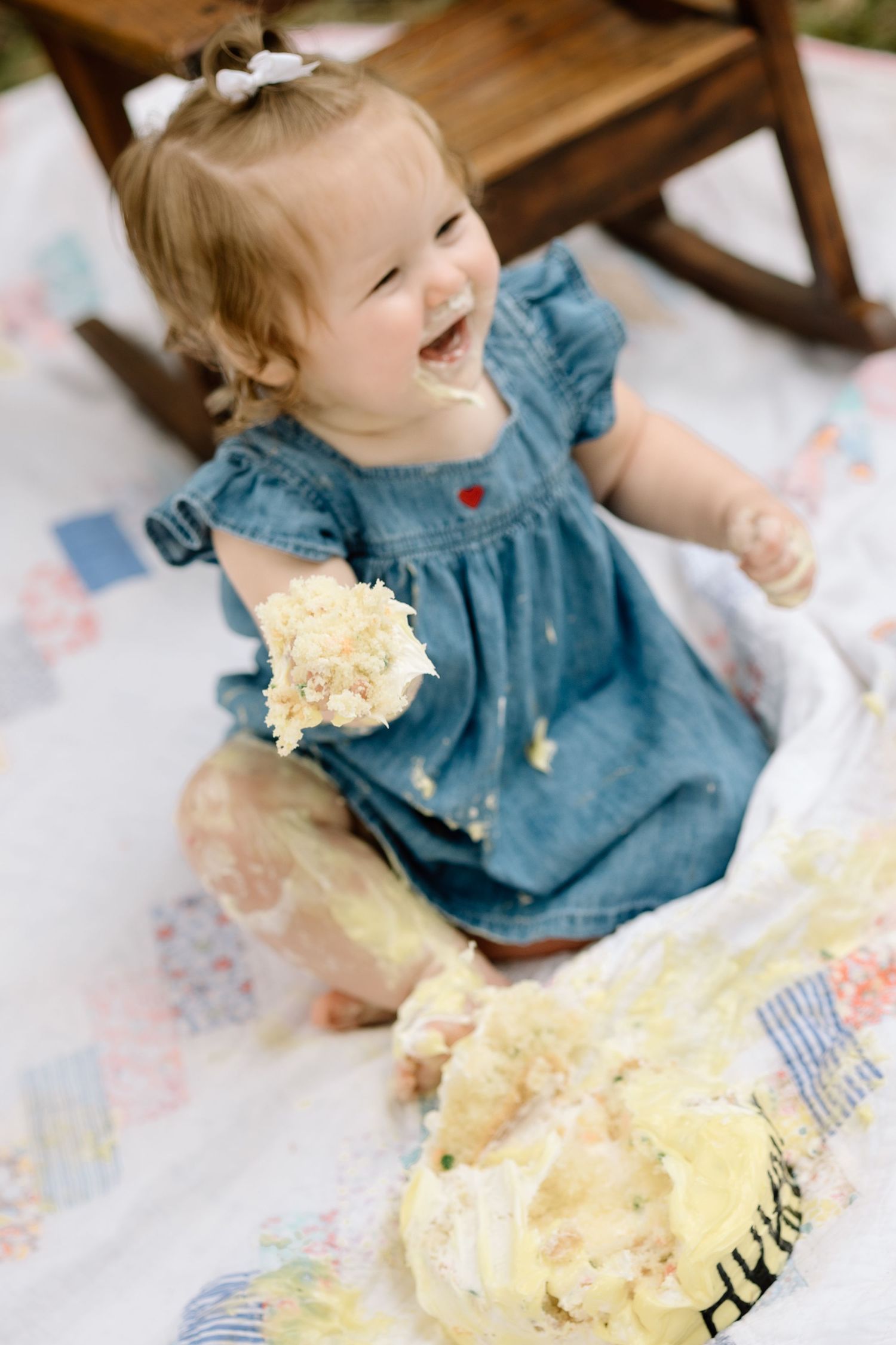 Joyful baby girl in denim dress holding smashed birthday cake during outdoor cake smash session.