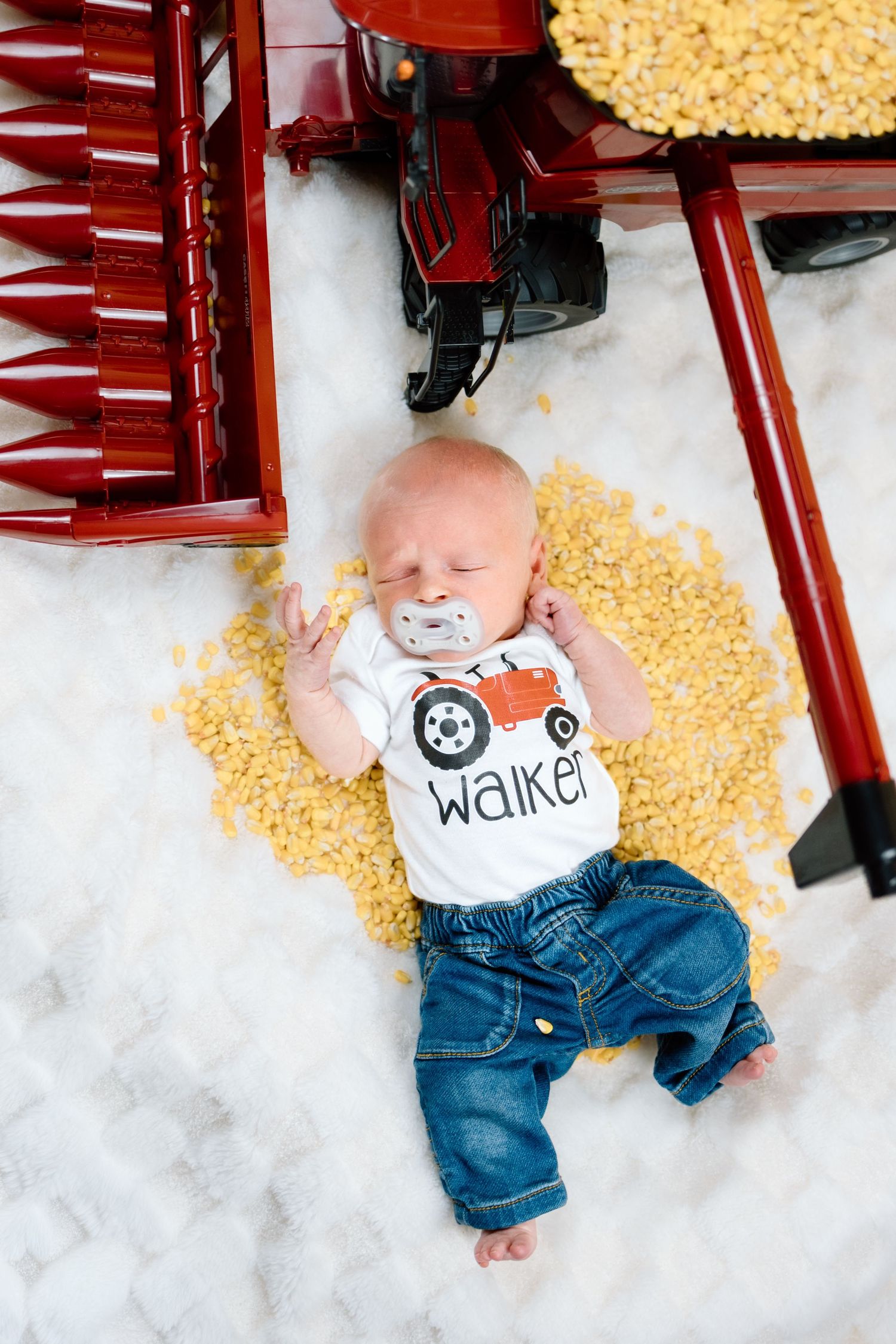 Newborn baby named Walker in tractor onesie and jeans sleeping among corn kernels beside large red toy farm equipment.