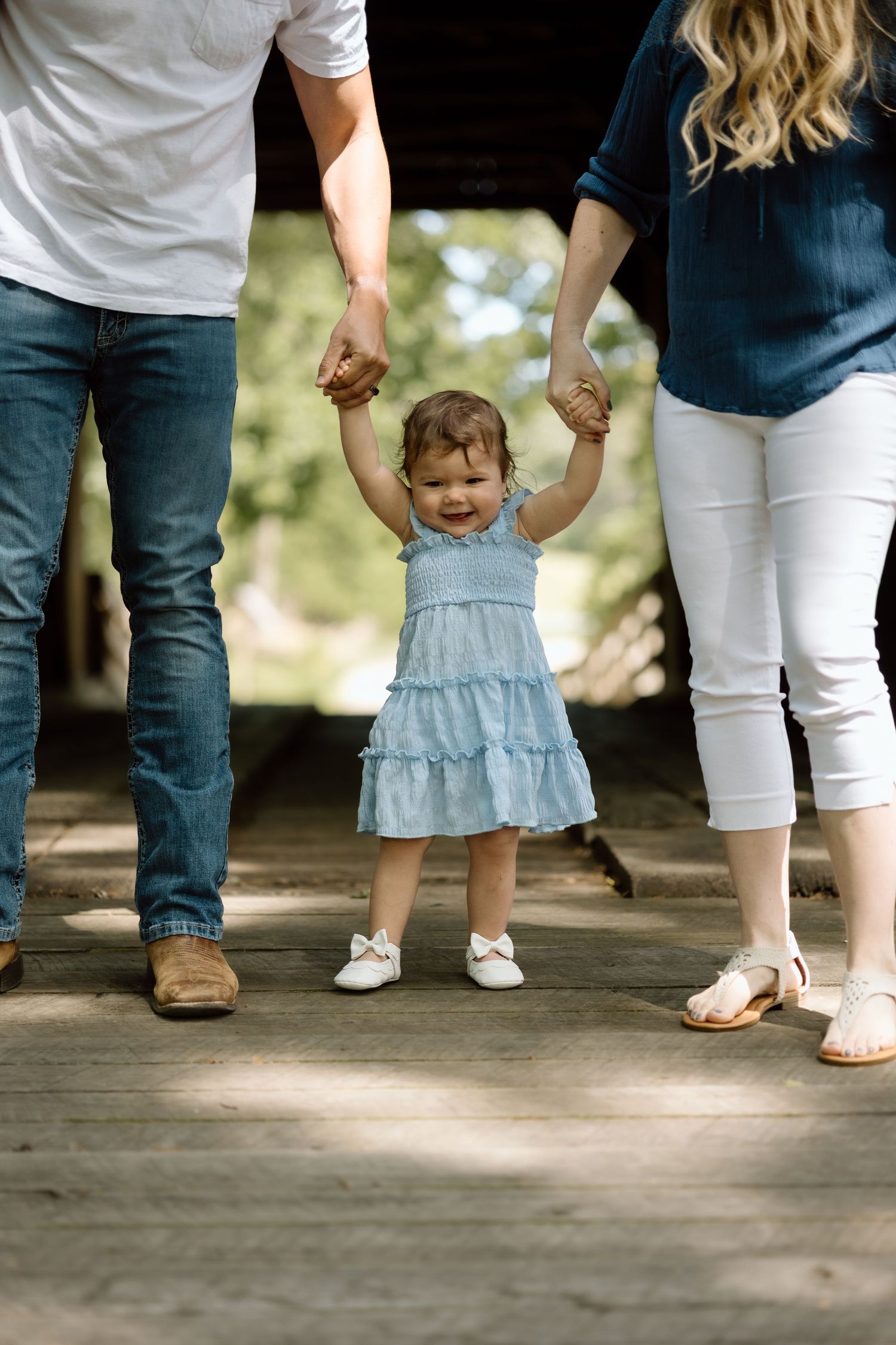 Toddler girl in blue dress holds parents hands while walking on wooden bridge during family photo session.