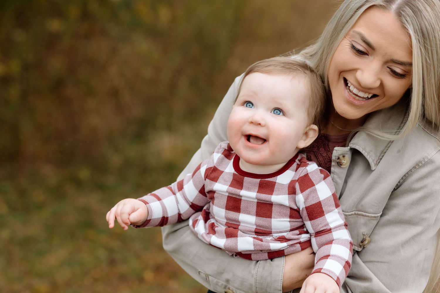 Blonde woman smiling warmly while holding a laughing baby in a red plaid shirt in an autumn setting.
